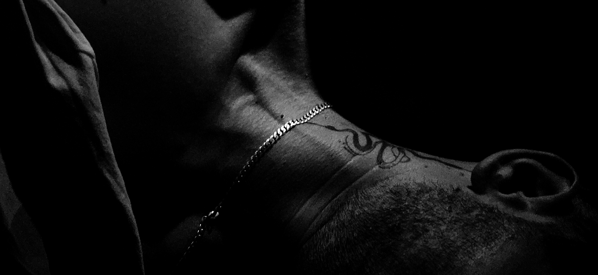 The black and white, low-key, tightly cropped image focuses on the neck, shoulder, and ear of an individual. The subject has a visible tattoo of what appears to be a stylized snake or knot on their neck. A silver chain necklace is worn around the neck, resting on the collarbone and extending out of frame. The lighting casts deep shadows, highlighting the contours of the neck and shoulder muscles. The background is entirely black, suggesting a dark or unlit environment. The individual's skin has visible texture, and stubble is present on the jawline and neck. The image is a close-up, abstract composition focusing on form, texture, and the play of light and shadow. No location in the city of Unknown, Unknown is discernible from the image.