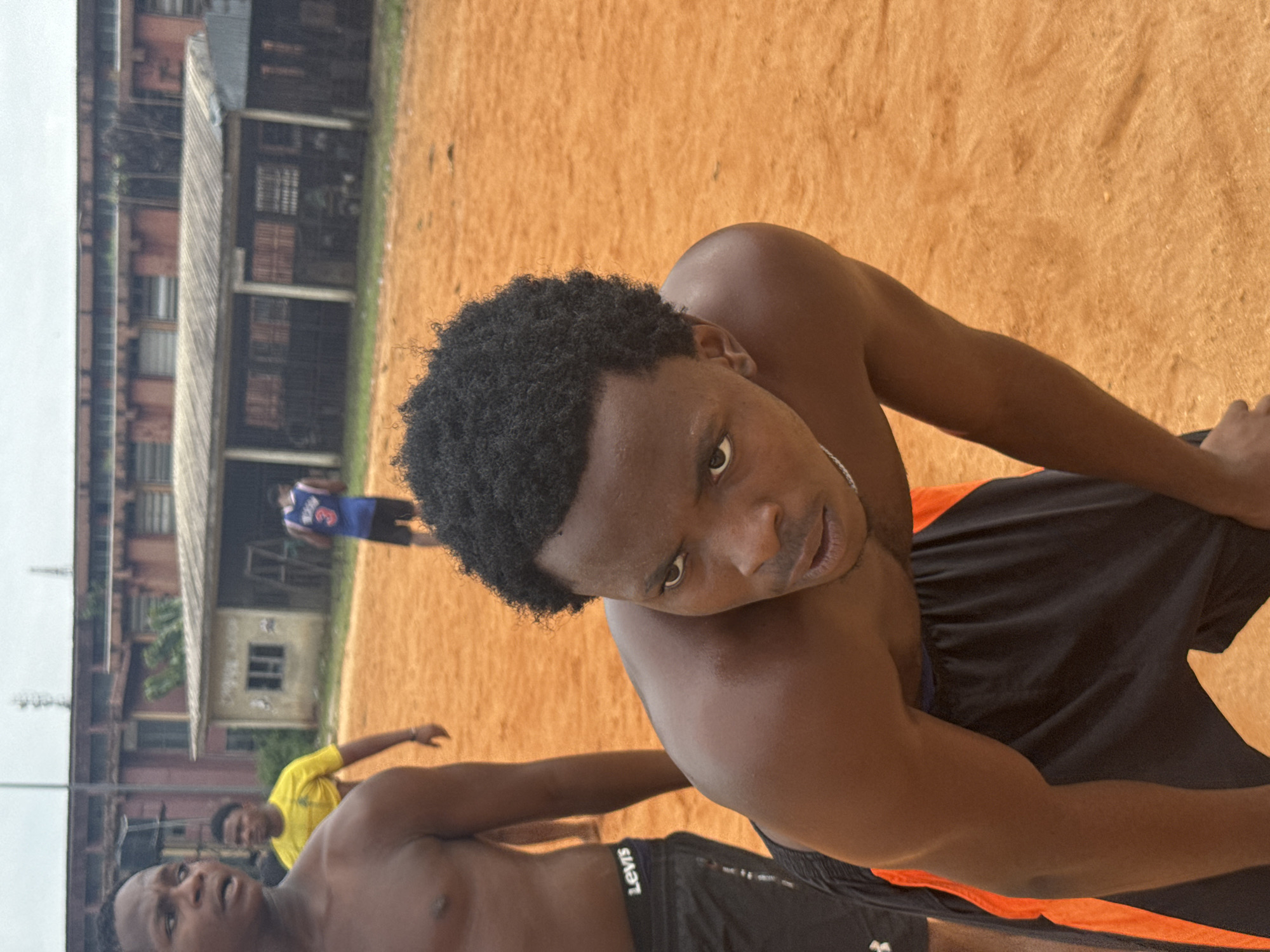 In Ihiagwa, Nigeria, this image captures an outdoor scene on a dusty, orange-colored court, likely a sports field. The focus is on a young Black man in the foreground, shirtless and wearing black shorts with orange trim. He is bent over, resting his hands on his knees, with a determined or exhausted expression on his face as he looks slightly off-camera to his right. His curly, dark hair is short. Behind him, to the left, another young Black man, also shirtless, is partially visible, his left arm extended. Further back and to the right, another individual wearing a blue basketball jersey with the number "3" and black shorts is also visible. In the background, a low-slung, weathered building with a corrugated metal roof and several windows or openings stands behind the court. A larger, older brick building with many windows is visible behind that, suggesting an urban or semi-urban setting. The overall atmosphere suggests a break during or after a sporting activity.