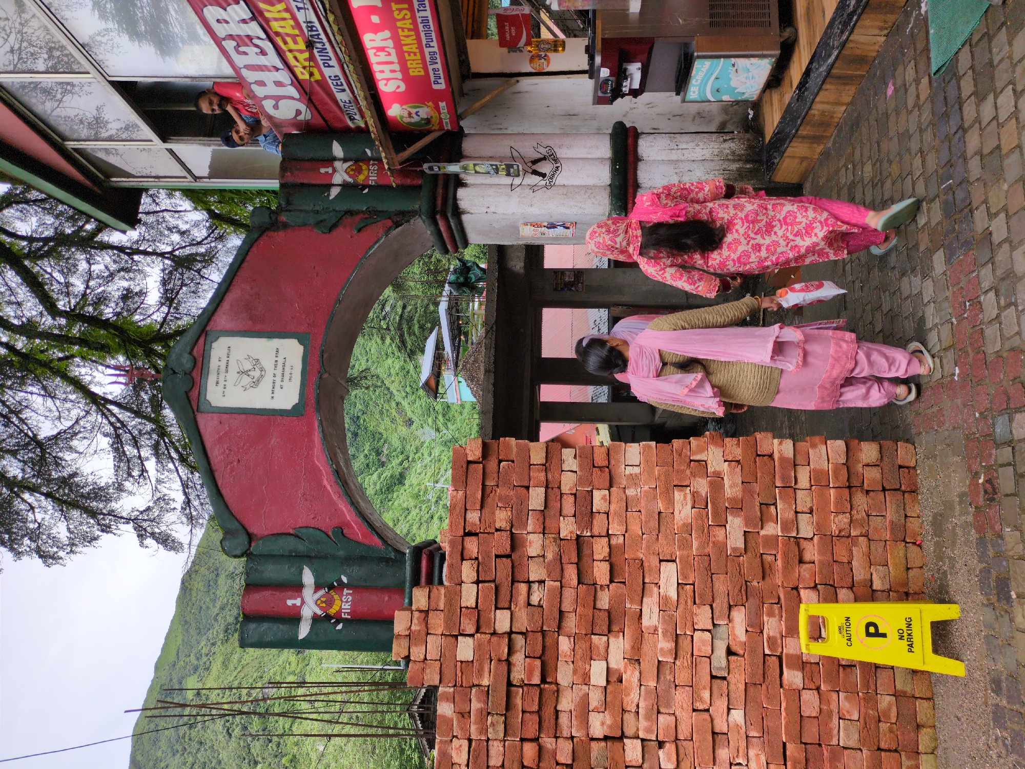 This image depicts the entrance to a building or establishment in Bhagsu, India. The entrance features a large, arched gateway painted in red and green, with decorative elements and a plaque bearing text. Two women, dressed in traditional Indian attire, are walking away from the camera, passing by a large stack of red bricks and a yellow "No Parking" sign. To the right of the arch, a shop or restaurant is visible, with signage indicating "SHER BREAKFAST" and "PURE VEG PUNJABI TASTE." Two men are looking out from a window on the upper floor of the establishment. The background reveals lush green hills and trees, suggesting a mountainous or hilly terrain. The ground is paved with stone tiles. The overall scene appears to be a bustling street or entrance area in a town or village setting.