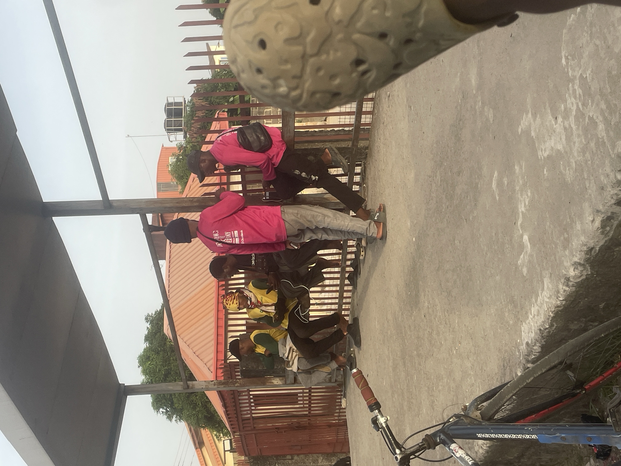 Four people are standing in front of a fence, leaning against it. The person closest to the camera is wearing a pink shirt and a black hat, with a black bag slung over his shoulder. Next to him is another person in a pink shirt, followed by a person in a yellow and green shirt, and finally another person in a yellow and green shirt. A bicycle is visible in the foreground. The location appears to be outdoors in Ado, Nigeria.