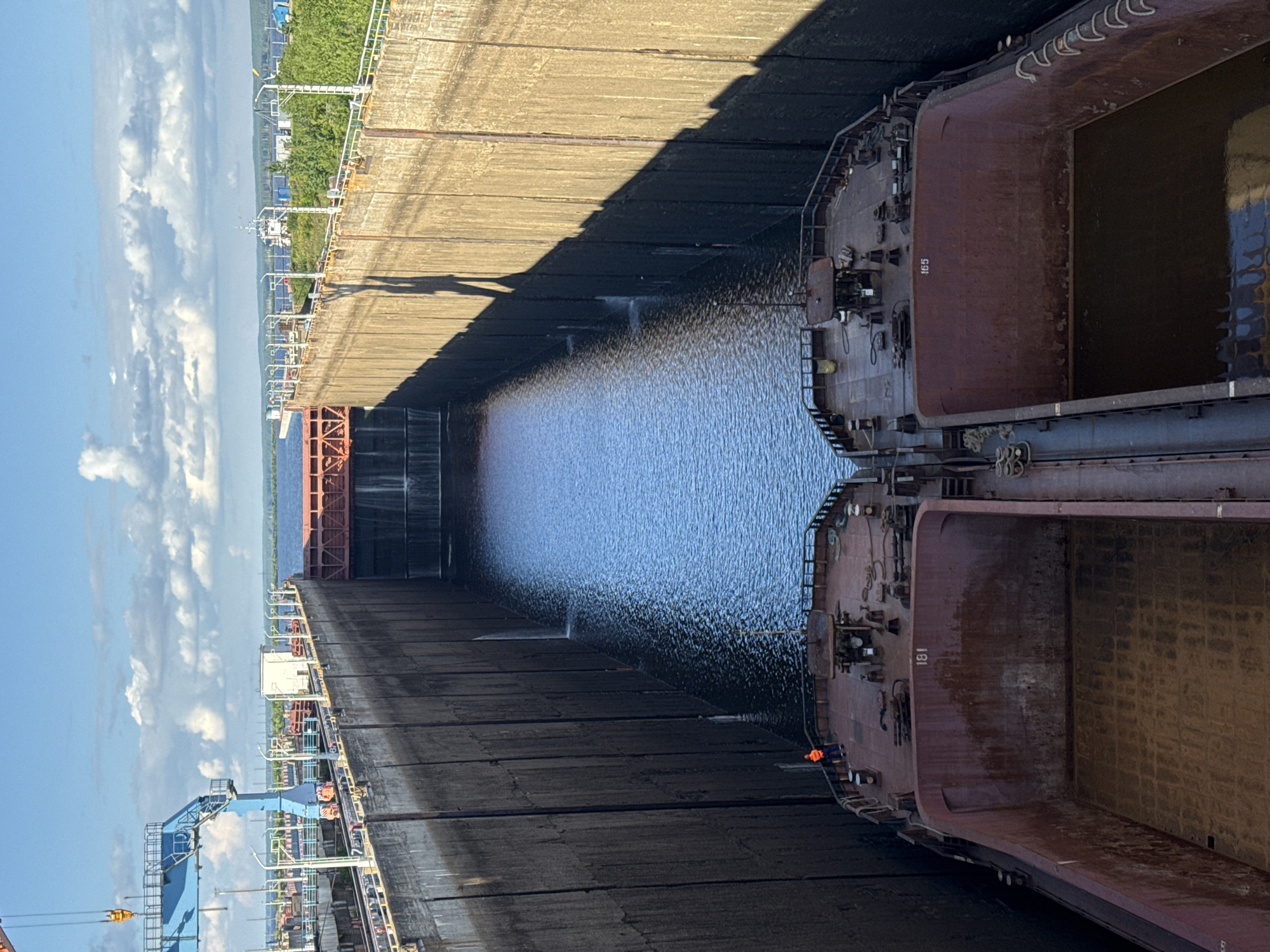 The image shows a view from above into a narrow, concrete-lined canal or lock, with two barges docked side-by-side at the bottom. The barges are empty and appear to be made of weathered red metal, with numbers "181" and "165" painted on their sides. A person wearing an orange vest is standing on the deck of the left barge. Water is flowing down the sides of the canal walls in several places, indicating that the lock mechanism is in operation. At the far end of the canal, a red metal gate is visible, with water cascading over it. The sky is blue with scattered white clouds, and sunlight casts long shadows on the canal walls. In the background, there are industrial structures, including a large blue crane on the left, and trees and water suggest a semi-industrial, waterfront environment. This scene is likely at the Chaikovsky Lock on the Kama River in Chaikovsky, Russia, a facility used for navigation and hydroelectric power generation.