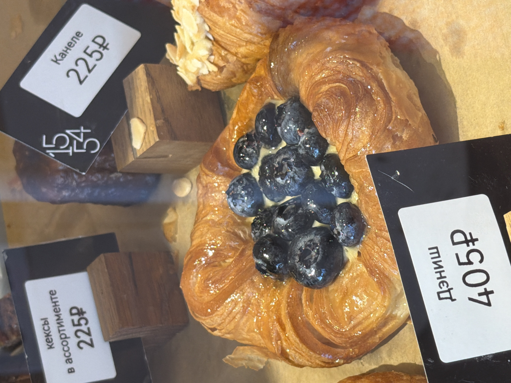 The image shows a display of pastries in what appears to be a bakery or cafe, possibly in Moscow, Russia. At least three types of pastries are visible with accompanying price tags. A blueberry Danish pastry is prominently featured, priced at 405 rubles. To the left, there are other pastries with labels "Канеле" (Canele) and "кексы в ассортименте" (assorted cakes), each priced at 225 rubles. Small wooden blocks are used as supports for the price tags. The image is taken from a close-up, slightly elevated angle, showing the arrangement of pastries in the display case.