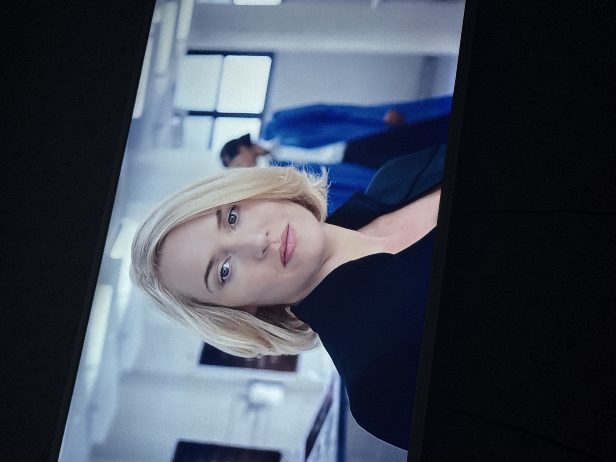 The image shows a woman with short blonde hair and a black top. She is looking at the camera. Behind her, there is another person in a blue garment. The background appears to be a modern, well-lit interior space, likely an office or studio. There is no visible evidence to determine a location in Mytishchi, Russia.