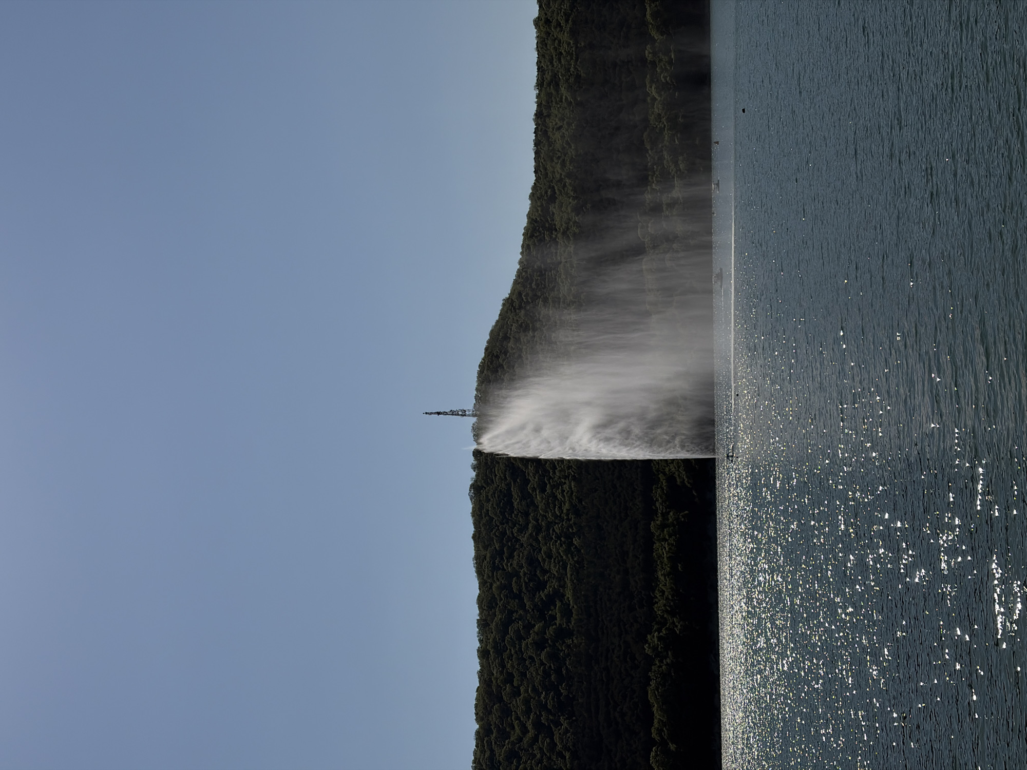 The image shows a large fountain in the lake, which is located in Abrau-Dyurso, Russia. The fountain appears to be spraying water high into the air, creating a misty effect. The background consists of a green, tree-covered mountain and a clear blue sky. The water in the foreground is sparkling and reflects the sunlight. The image is rotated 90 degrees counter-clockwise.