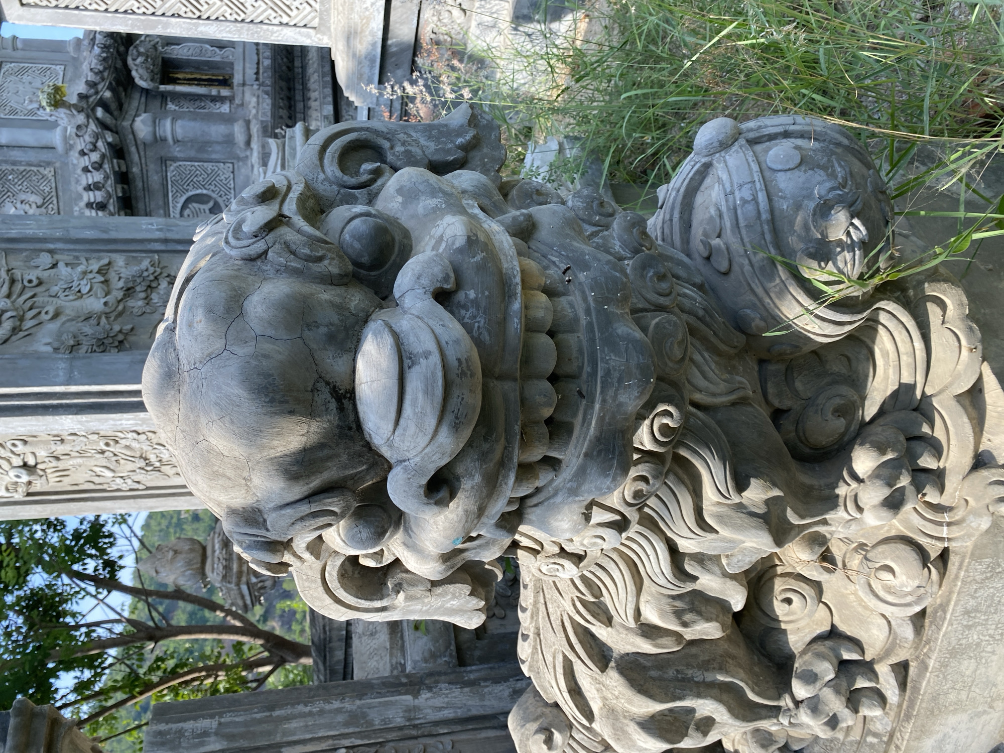 The image shows a close-up of a weathered stone statue of a mythical creature, possibly a Foo dog or a lion, in an outdoor setting. The statue is gray and appears to be made of concrete or carved stone, with visible cracks and signs of age. It has a fierce expression with bared teeth, large eyes, and ornate mane details. The creature is in a sitting or crouching position, resting its paws on a decorative base. In the background, there are other stone structures, possibly part of a temple or cemetery, and lush green vegetation, including tall grass and trees. The lighting suggests it is daytime, with sunlight casting shadows on the statue. The location is identified as Tay Nha Trang Ward, Vietnam. There are no people or visible actions in the image.