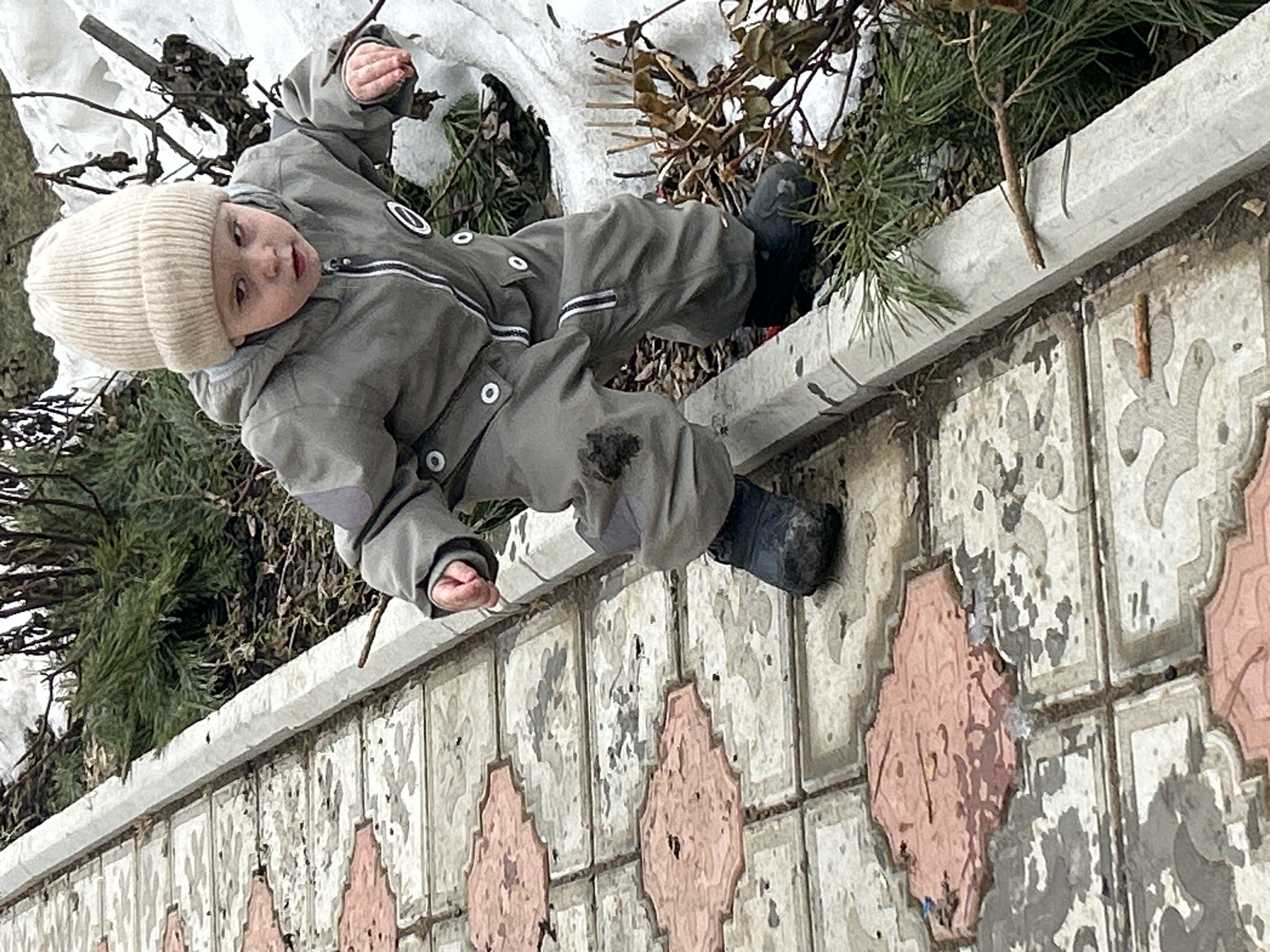 A young child wearing a beige knit cap and a gray snowsuit lies on a concrete ledge bordering a tiled surface in an outdoor environment that appears to be in Ольховка, Russia. The child's dark-colored shoes are planted on the tiles. Patches of snow and vegetation are visible behind the ledge. The tiles have a patterned design in shades of gray and pink. The scene suggests a cold or wintry season.