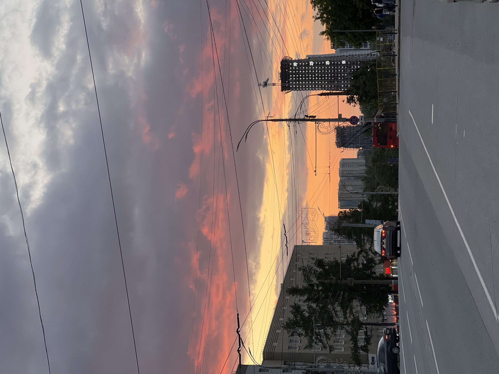 This is a vertical, eye-level shot of a city street at sunset in Izhevsk, Russia. The sky is filled with dramatic clouds painted in hues of orange, pink, and purple. Power lines crisscross the sky, and streetlights are visible. In the foreground, a multi-lane asphalt road stretches into the distance.
On the road, two cars are traveling away from the viewer. The car on the left is dark, and the car on the right is a lighter color with its brake lights illuminated. To the right of the road, a bright red trolleybus is visible, identified by the number "1413" on its side. Behind the trolleybus, a yellow barrier and several people standing and walking can be seen.
The cityscape features several multi-story buildings. On the left, a light-colored building with many windows is partially obscured by dark green trees. Further in the background, numerous modern apartment buildings and a tall, dark skyscraper under construction are visible. The skyscraper is illuminated, with numbers "15", "20", "25", and "30" displayed on its facade. The overall scene depicts a typical urban environment during a vibrant sunset.