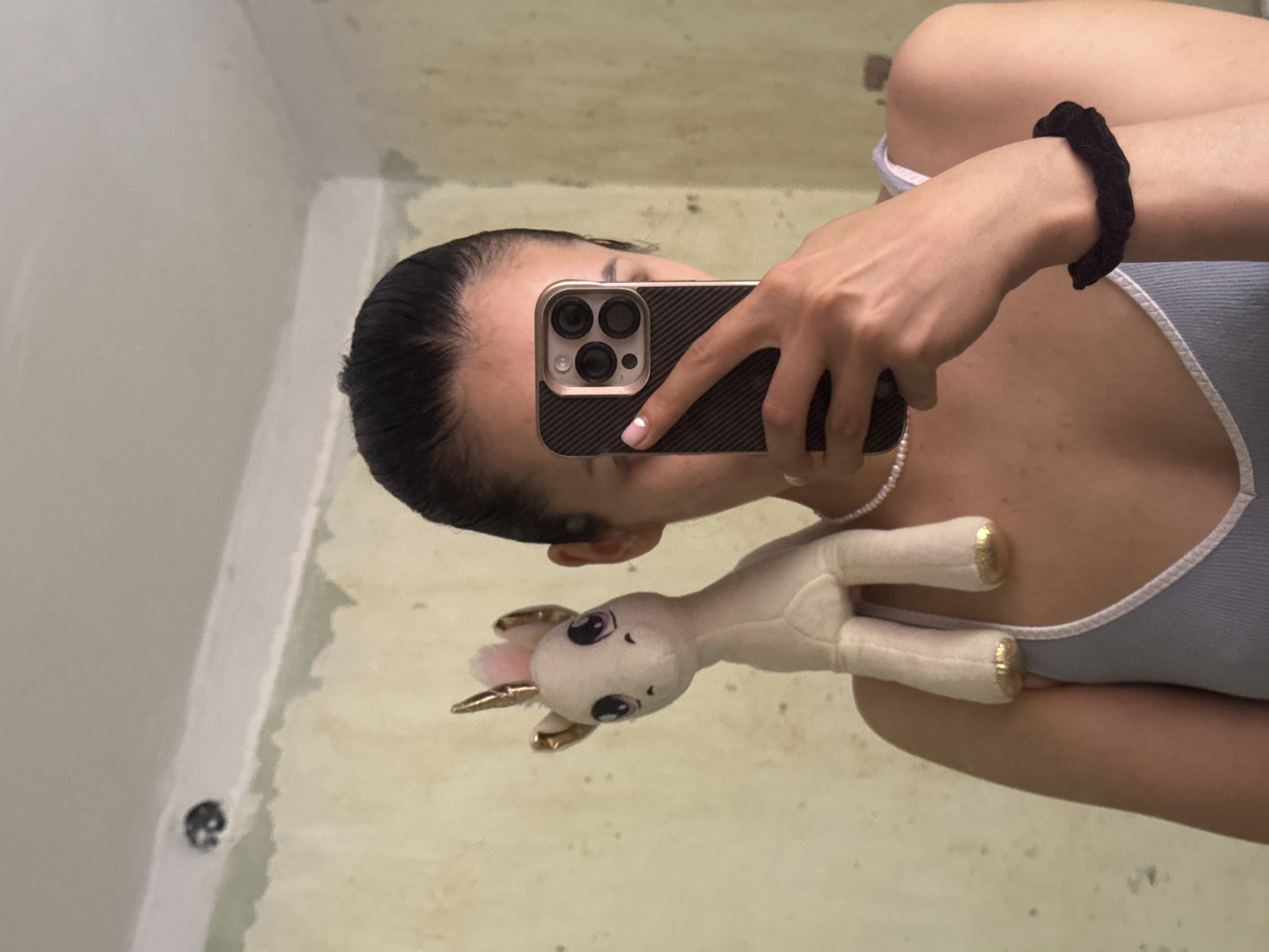 A young person is taking a mirror selfie in what appears to be a bathroom. They are holding a smartphone in their right hand, which is angled to capture their reflection and a small, plush unicorn toy. The person has their hair pulled back and is wearing a light blue tank top. A black scrunchie is visible on their left wrist. A pearl necklace is draped around their neck.
The background of the mirror shows a wall that appears unfinished or under renovation, with areas of exposed plaster and paint. There are some dark specks on the wall. The lighting is neutral, suggesting it could be daytime or artificial indoor lighting. The overall mood appears casual and playful, hinted at by the inclusion of the unicorn toy. No text is visible in the image. The location context is Votkinsk, Russia.