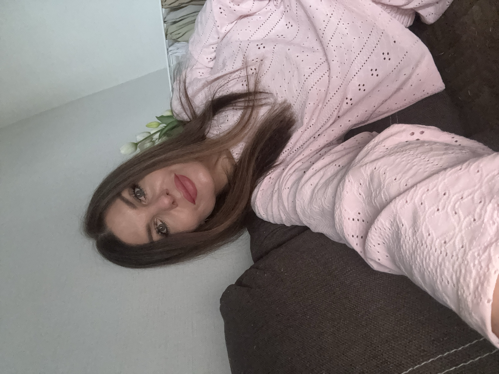 A woman with shoulder-length brown hair, wearing a pale pink eyelet-patterned top, is resting on a dark brown, textured couch. She is facing the camera with a slight smile and red lipstick. White flowers are partially visible near her head. The background consists of a light gray wall. The location is possibly Chaikovsky, Russia, but this cannot be verified.