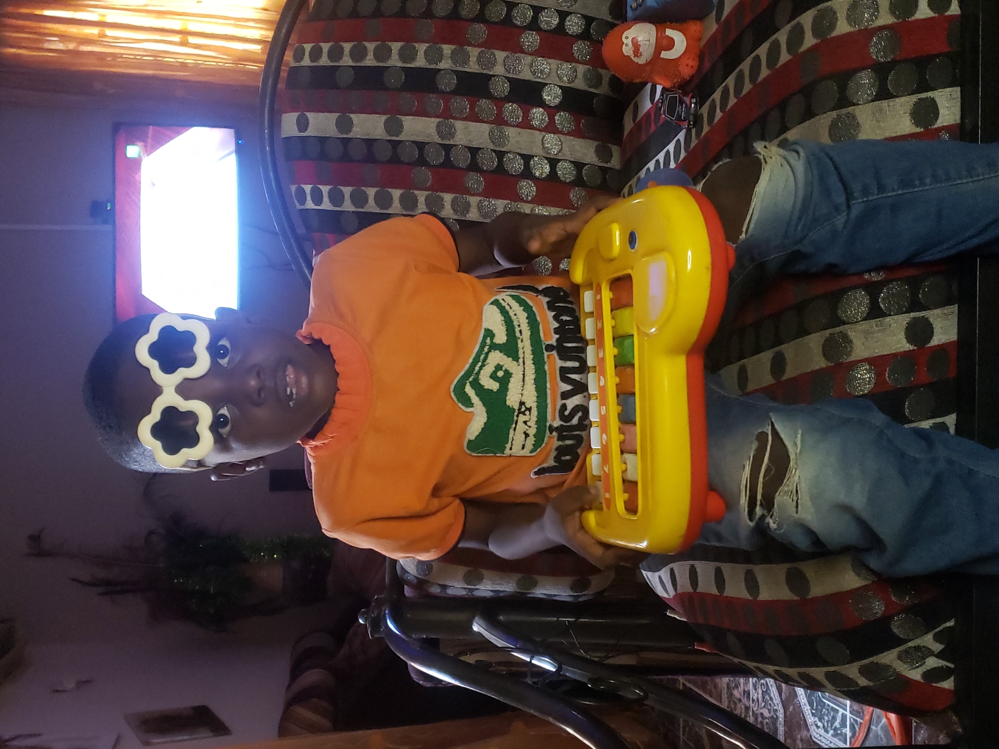 A young Black child is sitting on a patterned sofa and holding a yellow toy keyboard in a home environment in Sabon Tasha, Nigeria. The child is wearing an orange t-shirt with a graphic design and ripped blue jeans. The child has on white novelty glasses shaped like flowers over their eyes. The sofa has red, black, and white stripes with a pattern of silver circles. To the right of the child, on the sofa, are a small toy car and a plush orange monkey toy with white markings. In the background, a television screen is visible, displaying a brightly lit image, and a tall plant in a pot is to the left. The lighting in the room is dim.