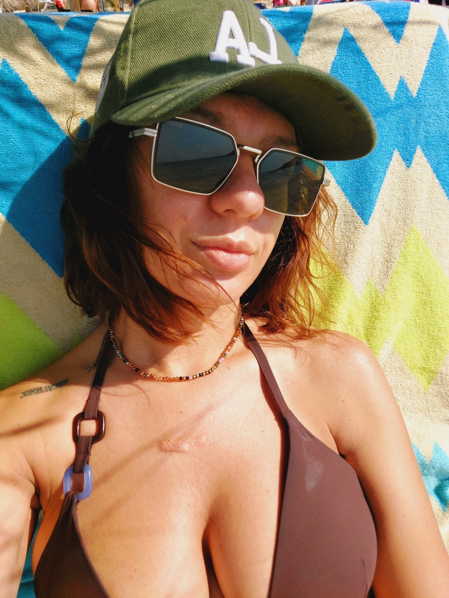 A woman wearing a brown bikini top and a green baseball cap is lying on a colorful beach towel. She has sunglasses on and a necklace with beads. A tattoo is visible on her left shoulder, and a scar is on her chest. The image appears to be taken outdoors, possibly at a beach in Sozopol, Bulgaria, with other people visible in the background.