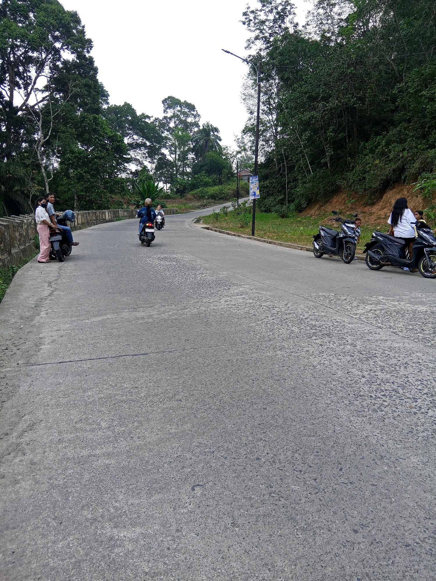On a slightly overcast day, an asphalt road curves uphill through a lush, green, and heavily wooded area. The road itself is rough and textured, with visible cracks and some wear. Along the left side of the road, a concrete barrier with decorative indentations separates the road from a lower, unpaved embankment.
At the start of the curve, on the left embankment, two people are standing near a parked black scooter. One person, wearing a white shirt and a light pink skirt, is looking towards the road. Next to them, a person in a light blue shirt and dark pants is standing, also looking away from the camera. A blue helmet rests on the seat of the scooter.
Further up the road, a person on a dark blue scooter is riding away from the camera. Another person on a lighter colored scooter is also visible further ahead on the road.
On the right side of the road, two black scooters are parked. The one in the foreground has a person with long black hair wearing a white shirt sitting on it, facing away from the camera. Beside them, a person in a yellow shirt is partially visible. A small child is peeking out from behind the second parked scooter.
A tall lamppost with a banner attached to it stands on the left side of the road, near the curve. The banner is blue and white with text and images on it. The surrounding environment is dominated by dense green foliage and trees, with some exposed soil on the hillside to the right. The sky is bright and overcast, suggesting diffused light. The scene appears to be in Lubuklinggau, Indonesia, given the presence of scooters commonly used for transportation and the tropical vegetation.