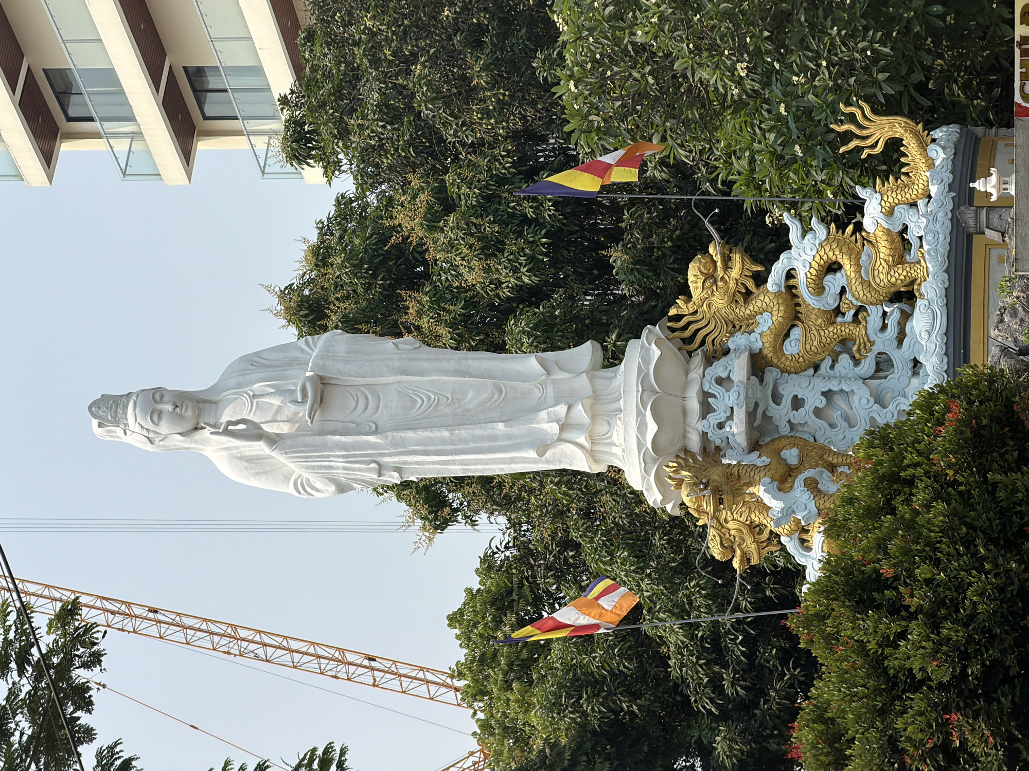 A large white statue of a woman is featured in this photograph, with an ornate gold and blue dragon sculpture behind it. The statue is positioned against a backdrop of lush green trees and foliage. A colorful Buddhist flag is visible on either side of the statue. In the background, the top of a multi-story building and a yellow construction crane can be seen. Based on the cultural symbols and architectural style, the location is likely in Đà Nẵng, Vietnam.