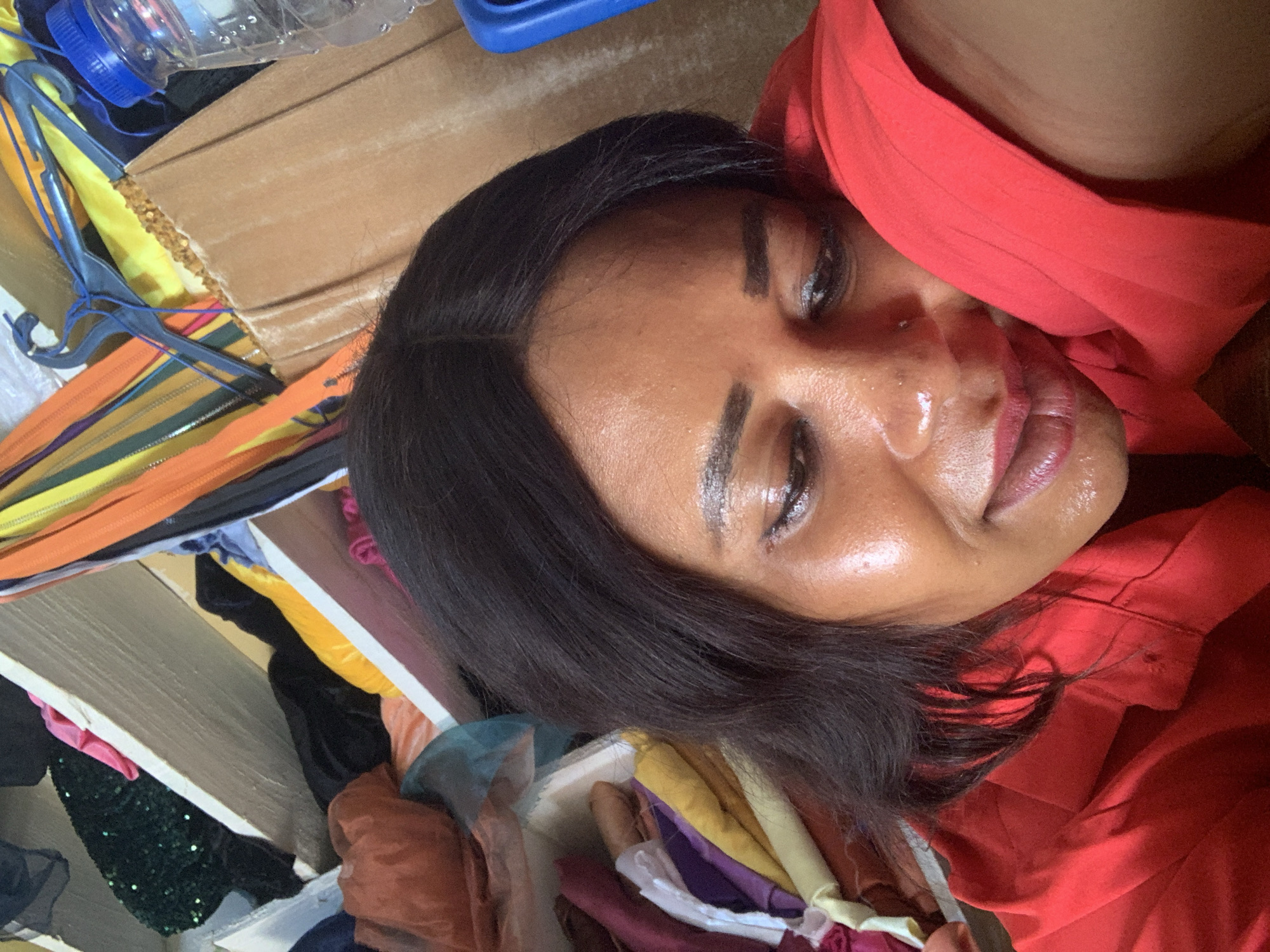 In Sabon Tasha, Nigeria, a close-up, indoor selfie captures a Black woman with dark hair, looking down and to the left, her left cheek resting on a bright red garment that appears to be a shirt. Her face is illuminated, highlighting a dewy complexion and subtle makeup. The background is cluttered with fabrics of various colors and textures, including what appear to be bolts of cloth, hangers, and a plastic water bottle, suggesting a sewing or crafting environment. The overall scene is intimate and personal, with the woman's expression conveying a sense of introspection or weariness.