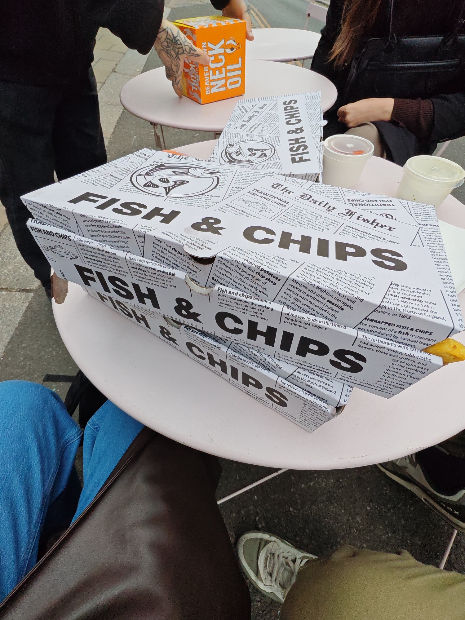 A high-angle shot depicts a casual outdoor dining scene, presumably in Reigate and Banstead, United Kingdom, given the style of the fish and chips packaging. Two light pink, round tables are visible, each hosting takeaway food containers. One table displays a distinctive orange box labeled "BEAVER NECK OIL." On both tables, containers are labeled "FISH & CHIPS" in a newspaper print design, along with white cups, possibly for condiments.
Three individuals are partially visible around the tables. One person wears a black outfit and sits to the right, their hand visible near the cups. Another person to the left wears black pants and has a tattooed hand near the "BEAVER NECK OIL" box. The third person is positioned lower in the frame, wearing blue jeans and a brown bag is draped over their lap. They also wear a grey sneaker.
The tables sit on a paved surface, suggesting an outdoor eating area in an urban setting. The focus is on the food packaging and the informal nature of the meal, rather than specific architectural details or landmarks.
