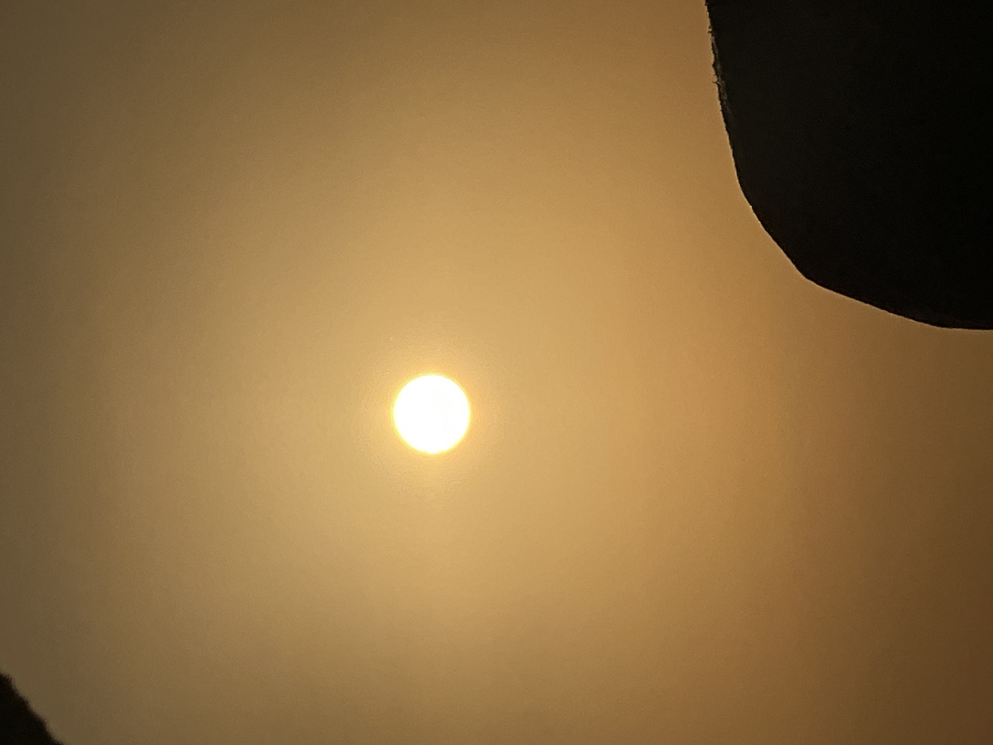 The image captures a bright sun, partially obscured by a dark, silhouetted object in the foreground, against a hazy, orange-toned sky. The sun appears as a luminous, circular disc. The silhouetted object, which occupies the lower right portion of the frame, has an uneven, rocky or mountainous texture. The overall scene suggests a sunset or sunrise, likely taken outdoors in a location that experiences atmospheric haze or pollution, which is common in bustling urban areas like Kuta, Indonesia, known for its beaches and vibrant atmosphere.