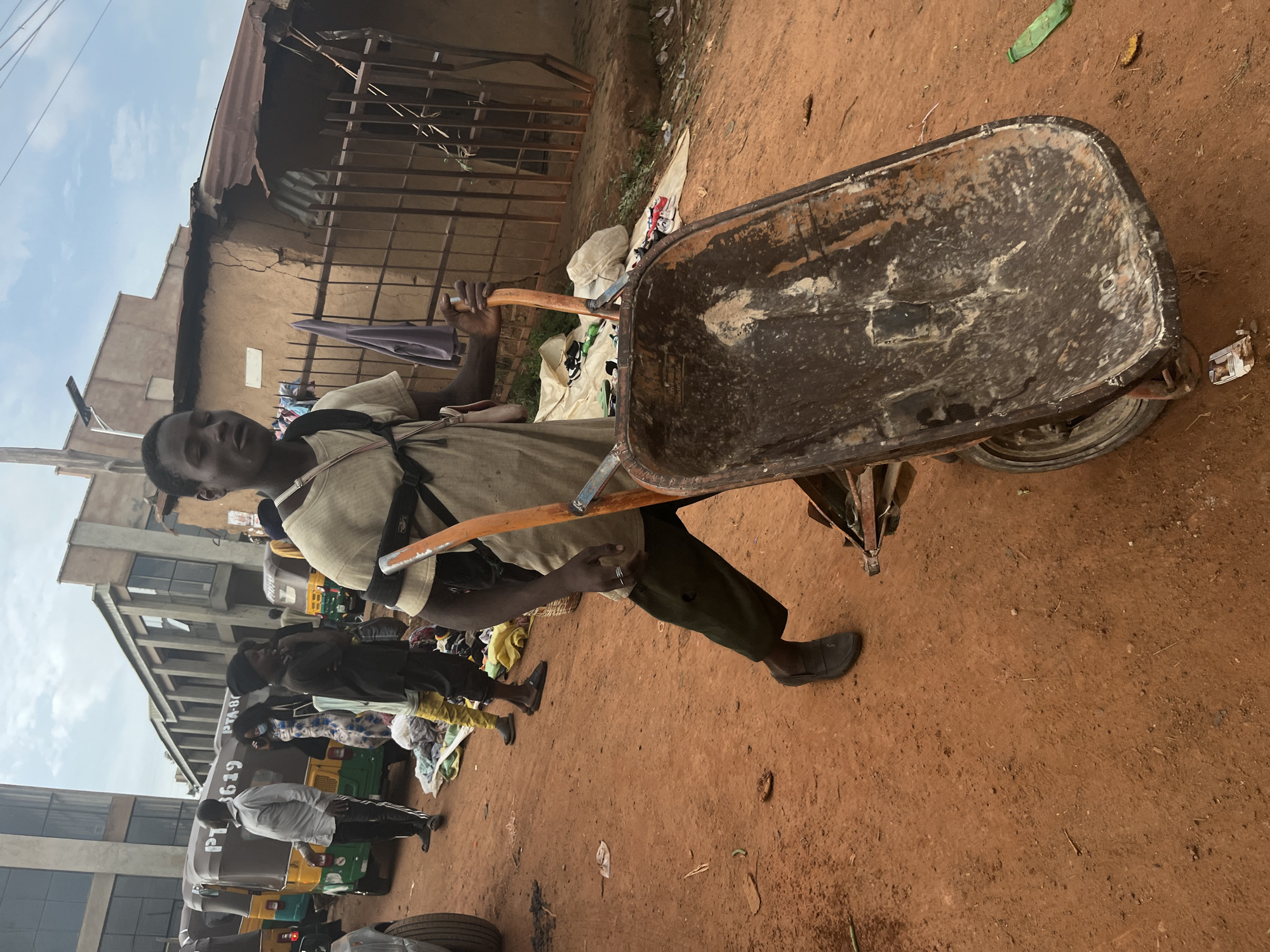 In Gura Top, Nigeria, a young Black male is pushing a rusty, orange-handled wheelbarrow filled with dirt on a reddish-brown dirt road. He is wearing a light tan short-sleeved shirt, dark pants, and dark shoes. He has a black backpack on and is holding the right handle of the wheelbarrow with his right hand. Behind him, several auto-rickshaws are parked. The rickshaws are yellow and green with "PTA-B19" and "PTA-B11" visible on them. Several other people are visible in the background, some walking and others standing. To the right of the road is a corrugated metal fence, a brown building with a tin roof, and some debris. The sky is overcast.