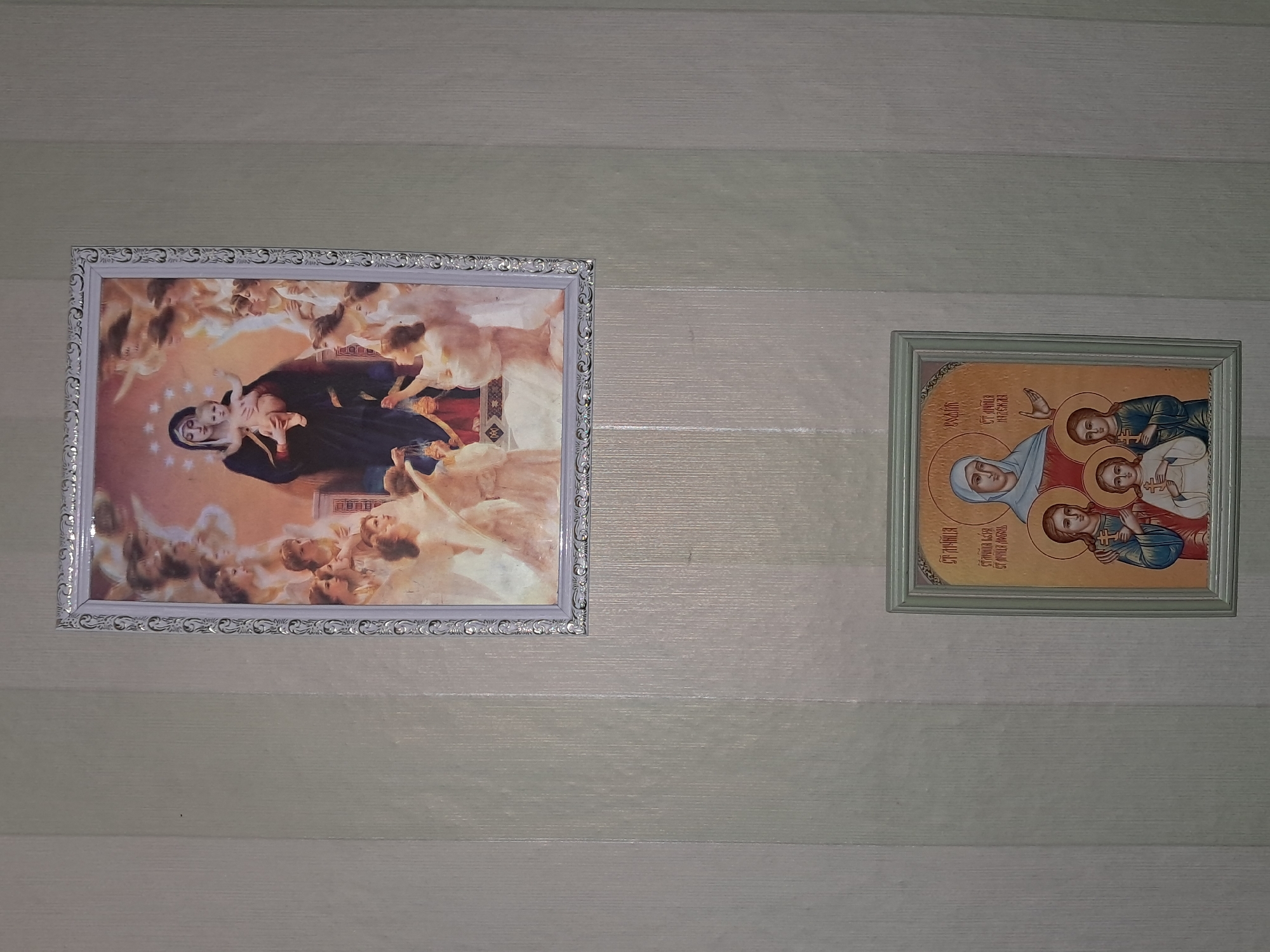 The image displays two framed religious icons hanging on a wall. The icon on the left is a large, rectangular painting in an ornate silver frame. It depicts the Virgin Mary holding the infant Jesus, surrounded by a multitude of cherubs or angels. The figures are rendered in a classical style with soft colors and dramatic lighting. The icon on the right is smaller and square, with a simple green frame. It features three figures with halos, characteristic of Eastern Orthodox iconography. The central figure, presumably the Virgin Mary, is shown with two other figures, likely Jesus and another saint. Both icons are depicted as being in a domestic setting, likely a home, with the wall behind them having a horizontal striped pattern. No specific time of day, weather, or strong emotional cues are discernible from the image. There is some Cyrillic text visible on the smaller icon, but it is too small and blurry to be read accurately. The location context of Shymkent, Kazakhstan, suggests these are likely Orthodox Christian religious artifacts.