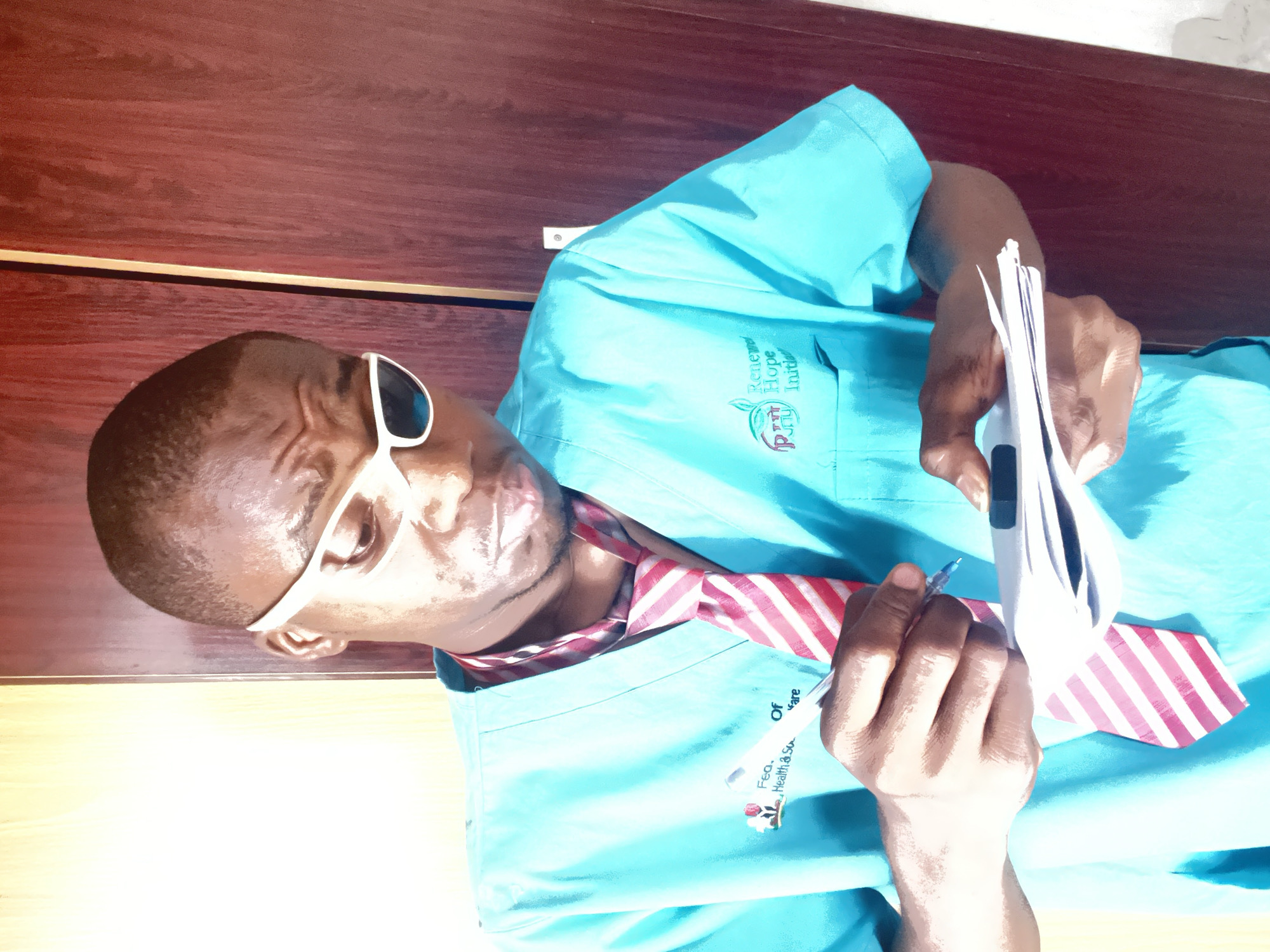 A man wearing white-framed sunglasses, a light blue medical scrub top, and a red and white striped tie stands in front of a dark wooden wall. He holds a stack of white papers in his left hand and a pen in his right, appearing to be writing. The medical scrub top has two logos on it. The first logo includes the words "Renewed Hope Initiative" and features a green apple and a red bird. The second logo includes the words "Fea. Health & So". The location of the image is not discernible as being in Minna, Nigeria, or anywhere else, based on the visual information.
