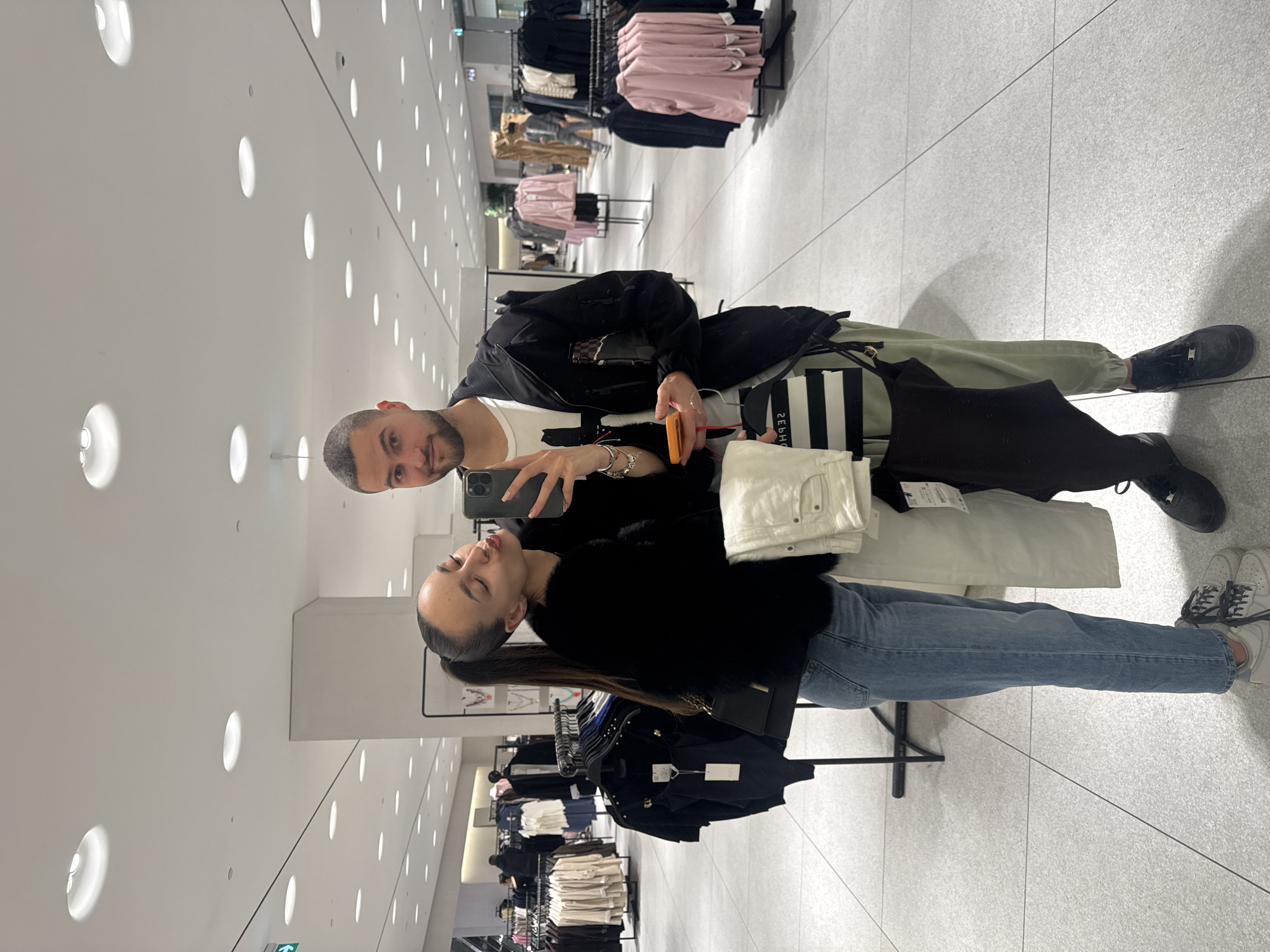 A mirror selfie depicts two individuals, a female and a male, inside a Zara clothing store in Paradise Center, Sofia, Bulgaria. The store features a modern interior with light grey large-format floor tiles and a white ceiling with numerous circular recessed light fixtures, some appearing as double units. A prominent white square column stands behind the couple.
The female, positioned on the left, has dark hair tied in a high ponytail. She wears a black faux-fur jacket, light blue jeans, and light grey sneakers. Her left arm is extended, holding a smartphone, as she puckers her lips for the photograph. A black shoulder bag hangs on her left side, and a ring is visible on her left ring finger.
The male, partially obscured on the right, has short dark hair and a beard. He wears a white t-shirt, a black zip-up jacket, light olive-green cargo pants, and black sneakers. He looks directly at the camera with a slight smile, holding the smartphone in his left hand and an orange item, possibly a phone case or small wallet, in his right. He also holds a folded pair of white trousers, a dark brown top, and a black-and-white striped Sephora shopping bag. Multiple bracelets are on his left wrist.
The background shows various clothing racks displaying garments such as light-colored tops, dark bodysuits, and pink blazers. Other shoppers are visible in the distant background. An emergency exit sign with a green arrow is located in the upper left corner of the reflection.