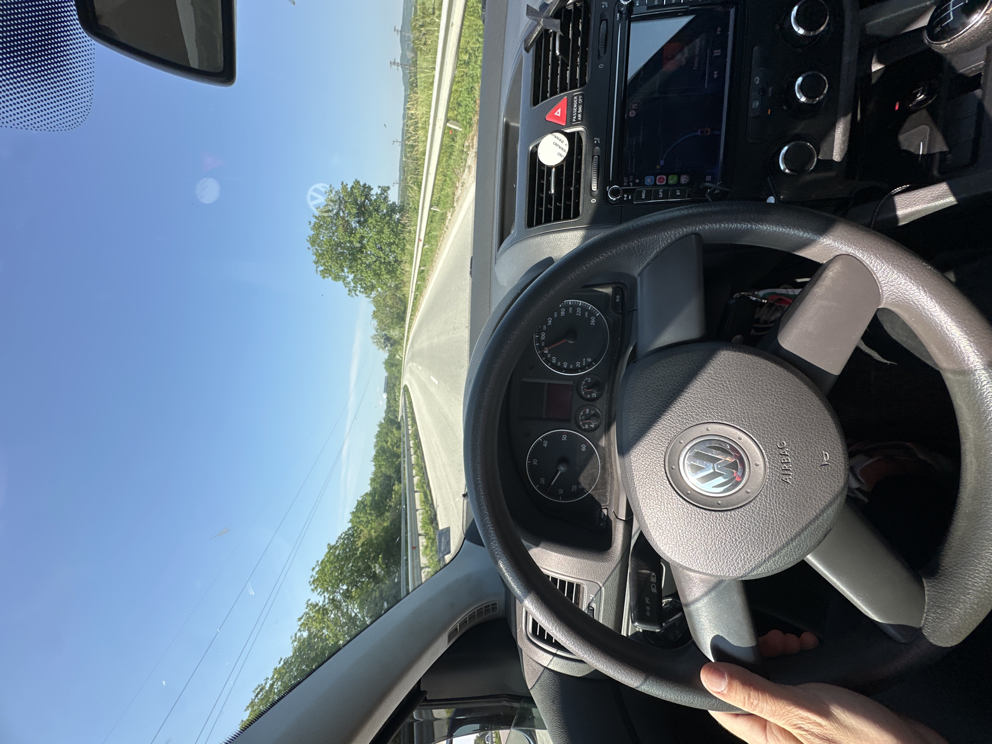 The view from inside a Volkswagen vehicle shows a highway with green vegetation on either side, under a clear blue sky. The dashboard instruments and a navigation screen are visible, along with the steering wheel. The speedometer indicates a speed of 50 km/h. A hand is gripping the lower right portion of the steering wheel. The scene may be on a highway near Lovech, Bulgaria.