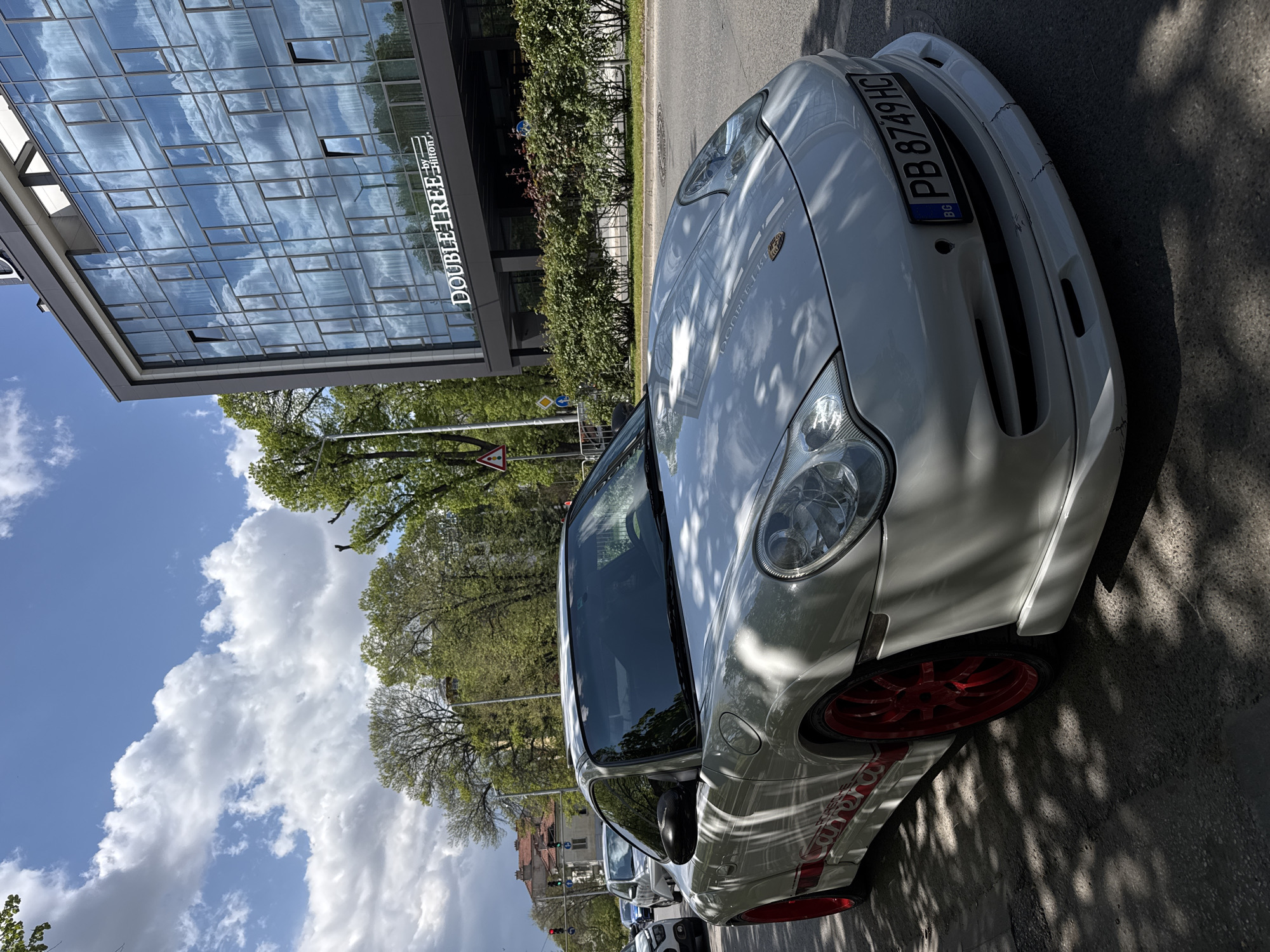 The image shows a white Porsche 911 parked on a street in Plovdiv, Bulgaria, facing the viewer’s right. The car has red rims and the license plate reads "PB 8749 HC." Behind the car, a DoubleTree by Hilton hotel building is visible, its glass facade reflecting the blue sky and white clouds. There are trees and other vegetation between the car and the hotel. In the background, other buildings and street signs are seen. The weather appears to be sunny with a partly cloudy sky.