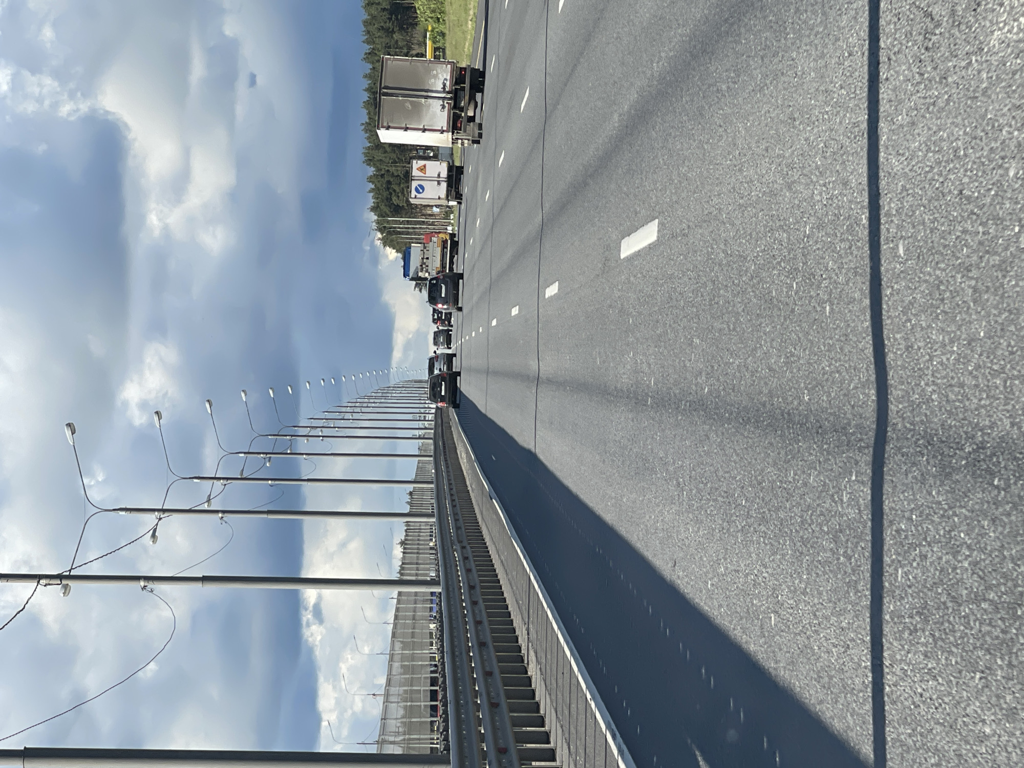 The image depicts a multi-lane highway, likely located in or near Slashchevo, Russia, as inferred from the surroundings. A line of vehicles, including trucks and cars, is visible in the distance on the left lanes of the highway. A metal barrier separates the left and right lanes. A series of tall streetlights line the side of the road. The road surface appears to be asphalt with lane markings. The sky is partly cloudy. The scene is a view from a vehicle traveling on the right side of the highway.