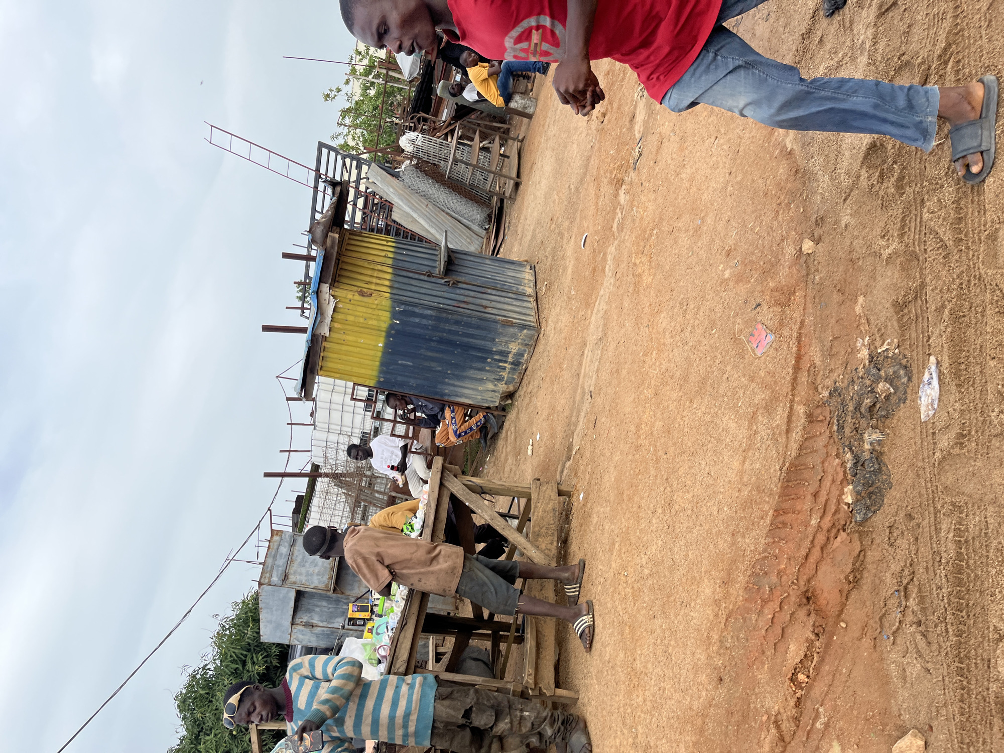 This image depicts a bustling outdoor market scene in Gura Top, Nigeria. Several individuals are present, engaged in various activities. In the foreground, a person wearing a striped sweater and sunglasses is partially visible, holding a clear plastic bottle. Behind them, another person in a brown t-shirt and shorts stands near a wooden stall laden with goods, including what appear to be electronic devices.
Further into the background, other people are seated or standing around market stalls. One stall is constructed from corrugated metal, painted yellow and blue, suggesting it might be a more permanent structure within the informal market. A ladder is visible attached to scaffolding in the background, hinting at ongoing construction or repairs in the vicinity. The ground is sandy and appears well-trodden, with tire tracks visible in some areas. The sky is overcast, suggesting a cloudy or potentially rainy day. The overall impression is one of a lively commercial environment, with people actively participating in the day's trade.
