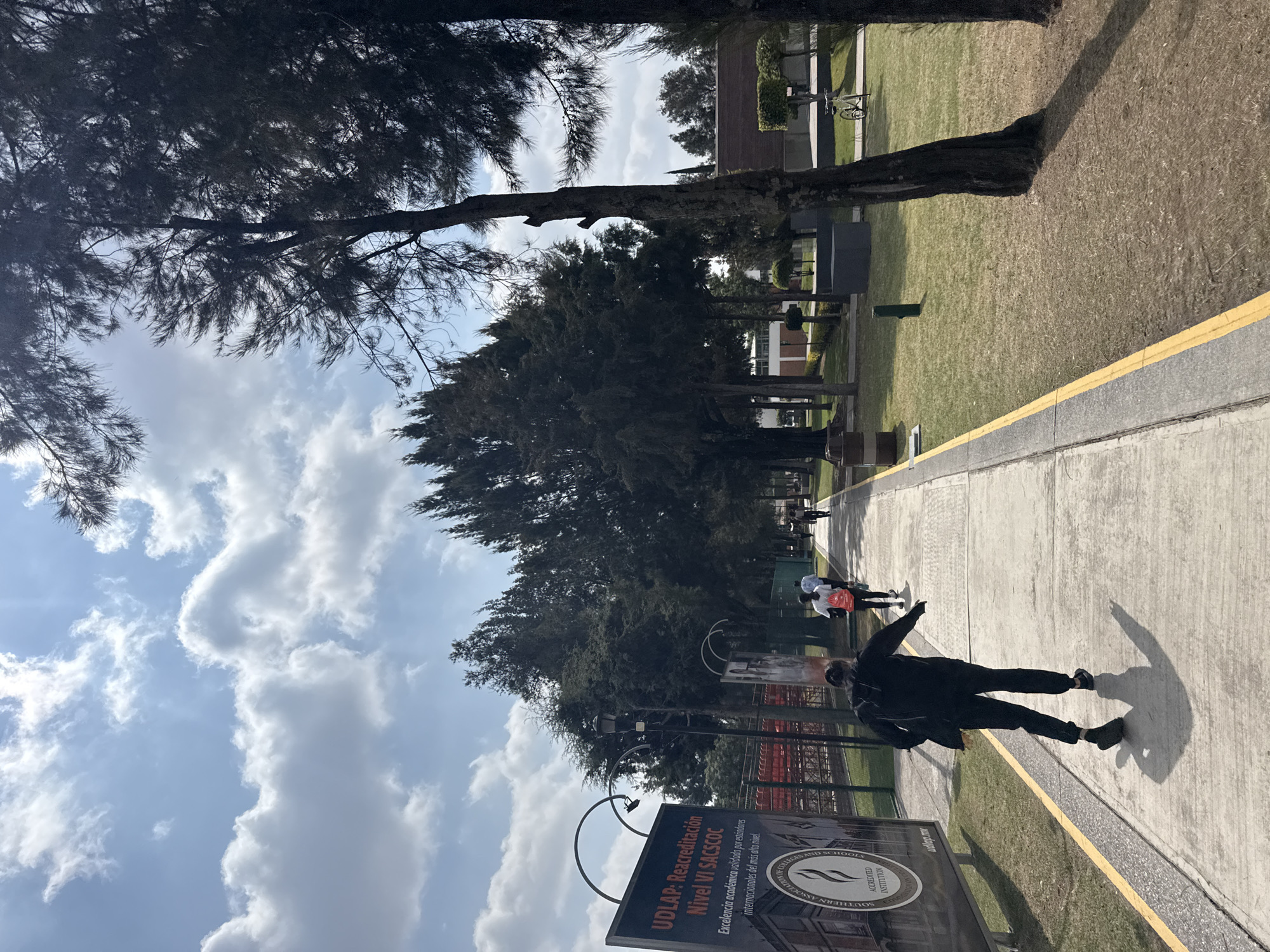 The image depicts a scene at the Universidad de las Américas Puebla (UDLAP) in San Andrés Cholula, Mexico. The view is from the sidewalk, where a person in dark clothing is walking with arms outstretched, casting a shadow. In the background, other individuals are walking along the path. The scene includes trees, grass, and a sign displaying UDLAP's accreditation level. The sky is visible with scattered clouds.