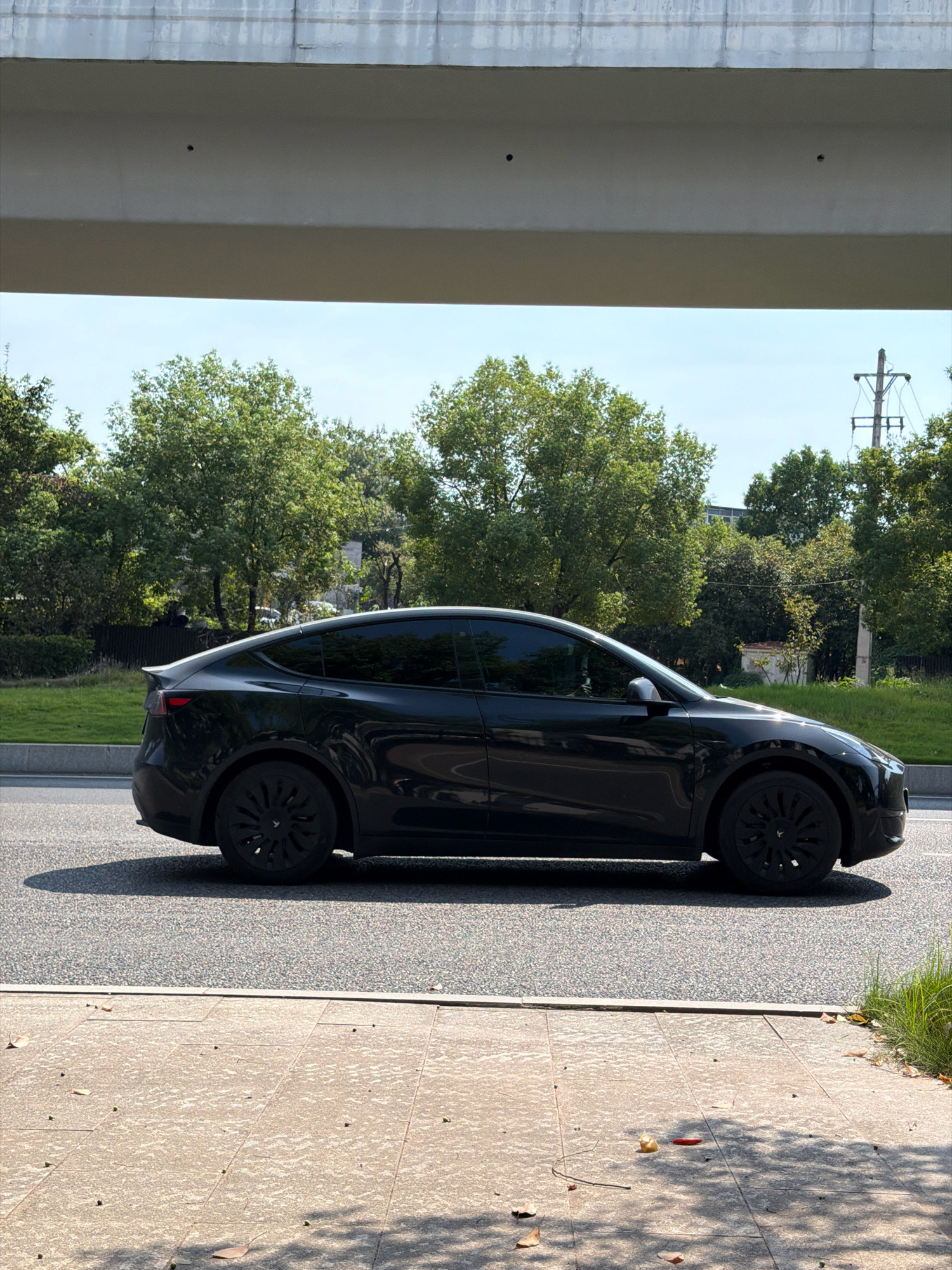 A black Tesla Model Y is parked on the side of a road in Jiangjin, China. The car is facing right and its side is visible. The road is paved with asphalt, and a sidewalk is to the right of the road, paved with rectangular stones. The car is parked on the left side of the road, with a grassy area and trees behind it. Above the road, there is a concrete overpass. The sky is blue with some clouds. There are no people visible in the image.