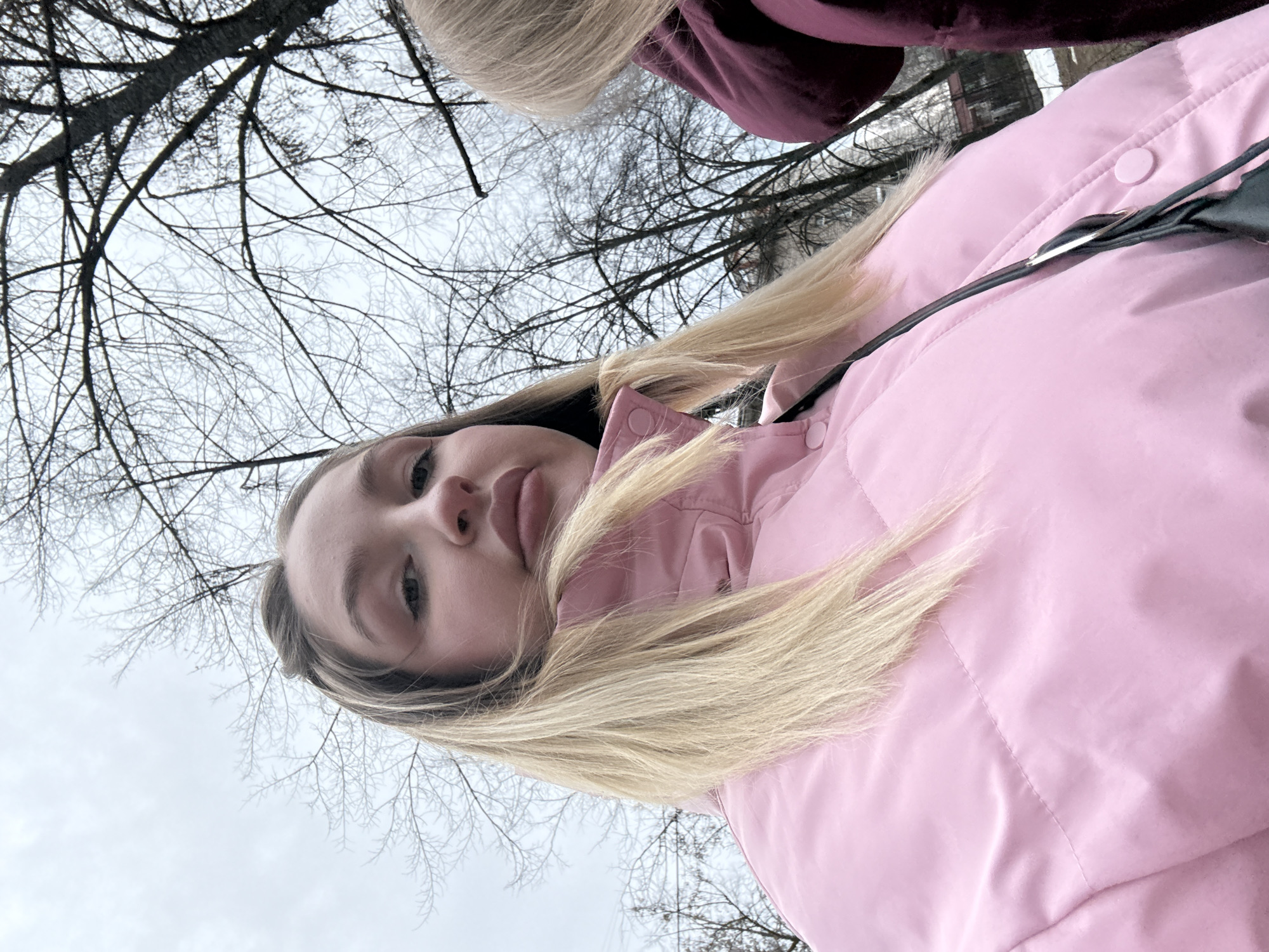 The image is a low-angle selfie of a young woman with light blonde hair. She is wearing a light pink puffer jacket. Part of another person wearing a dark burgundy garment is visible in the upper right corner. Behind the woman and the other person, bare tree branches are silhouetted against an overcast sky. The scene is outdoors, but the specific location in Chaikovsky, Russia, cannot be determined from the visual information provided.