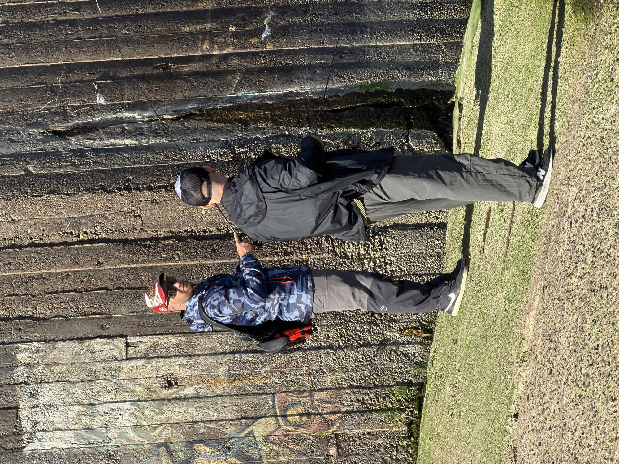 Two men are standing on a structure, which appears to be a bridge or wall, near what looks like a body of water, in Chaikovsky, Russia. The structure consists of horizontal, concrete-like layers. One man is wearing a blue camouflage jacket, gray pants, and a red and white hat. He is pointing his finger in the direction of the water. The other man is wearing a black jacket, gray pants, and a black baseball cap. He is holding a fishing rod. The structure they are on has grass growing on one side. There's possible graffiti on the side of the wall.