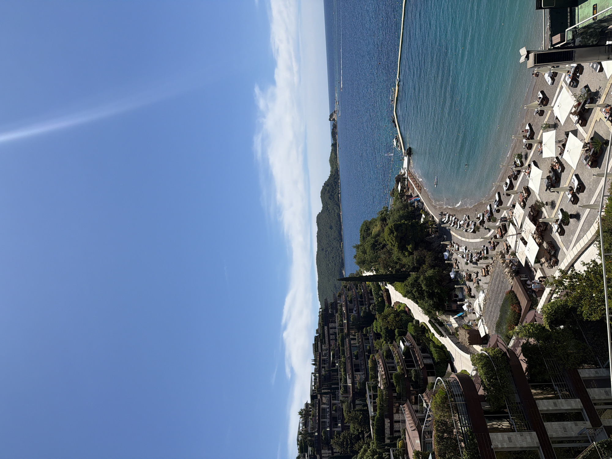 This is a high-angle, full shot of a coastal resort area in Budva, Montenegro, on a clear, sunny day. The foreground features a modern, multi-tiered hotel complex with balconies and lush greenery. A winding pathway leads down to a pebble beach lined with rows of lounge chairs and umbrellas, some occupied by people. Adjacent to the beach is a swimming pool area and what appears to be a restaurant or bar. The turquoise sea stretches out to the horizon, with a long pier extending into the water. In the distance, a verdant, tree-covered hill forms the backdrop. The sky is a brilliant blue with scattered white clouds.