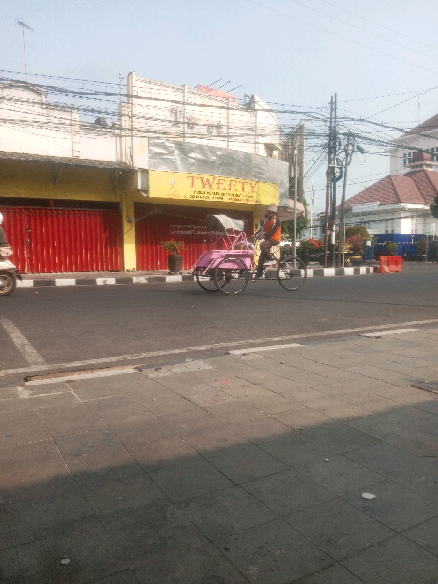 An outdoor, eye-level, medium shot captures a street scene in the city of Kediri, Indonesia. The foreground features a paved sidewalk with distinct squares, partially shaded by a warm, late afternoon sun. The street itself is asphalt, with white road markings and a curb.
In the mid-ground, a person on a pink and white bicycle rickshaw is moving from left to right across the frame. The rickshaw has a covered passenger area and two large wheels at the back and one at the front, all spoked. The driver wears a hat and an orange vest over a dark shirt and pants.
To the left of the rickshaw, a building with a bright yellow facade is visible, featuring large red roll-up doors. A sign above the doors reads "TWEETY" in large letters, with smaller text below indicating "PUSAT PERLENGKAPAN BAYI & ANAK JL. DOHO NO.02 - KEDIRI - TERLENGKAP DAN MURAH".
Further back, a building with a red roof is visible, and beyond that, another building with a white facade and a prominent clock tower. The sky above is clear and pale blue, with a complex network of overhead electrical wires crisscrossing the scene. A few potted plants add a touch of greenery to the urban landscape. The overall impression is of a typical street scene in an Indonesian city on a sunny day.