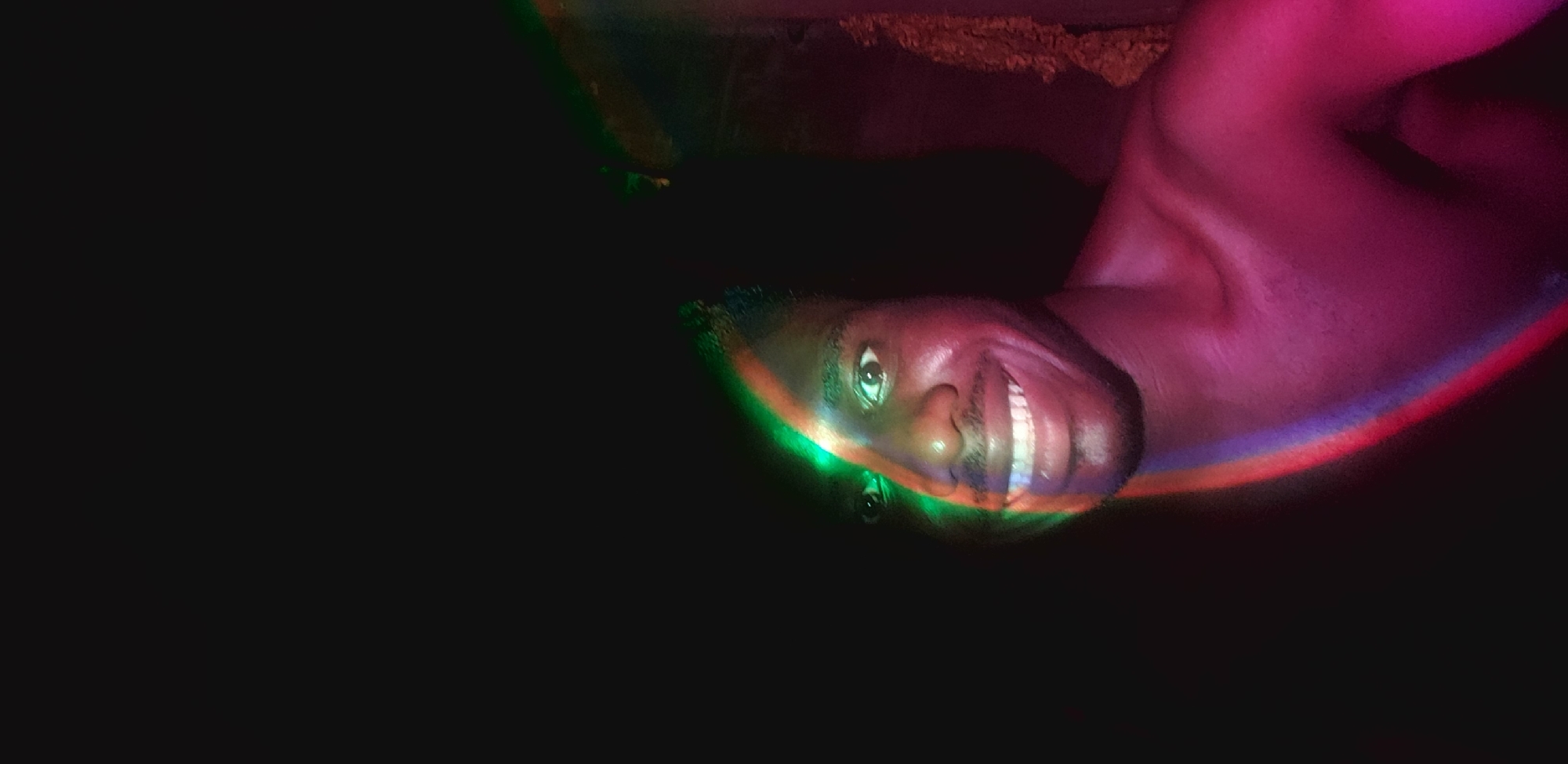 The image is a close-up, vertical shot of a Black man's face and upper chest, taken in what appears to be a dimly lit indoor environment. The man is looking directly at the camera with a wide smile, his teeth clearly visible. He has dark hair and a short beard. His skin is illuminated by a colorful light that appears to be a prism effect, casting green and red hues across the left side of his face and a pinkish glow on his chest and right shoulder. The background is predominantly dark, suggesting a low-light setting. There is some textured, reddish-brown material visible on the far right, possibly part of the wall or furniture. The image does not contain any information to indicate a location in Narayi, Nigeria.