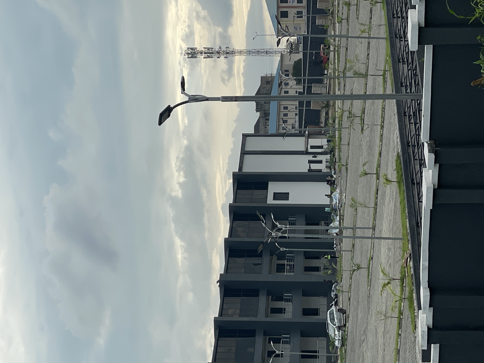 The image depicts a cloudy sky over a modern construction site in Abuja, Nigeria. The foreground features a dark grey wall with white trim, beyond which is a paved parking lot dotted with young palm trees. Several streetlights are strategically placed throughout the lot. In the mid-ground, a multi-story building with dark tinted windows and a grey facade dominates the scene. Construction activity is visible, with people working and materials laid out on tarps in front of the building. A silver car is parked to the left. Further in the background, a cell tower rises above other buildings, one of which has a blue roof, and a glimpse of another building with an advertisement is visible. The overall impression is that of ongoing development and urban expansion in a contemporary setting.