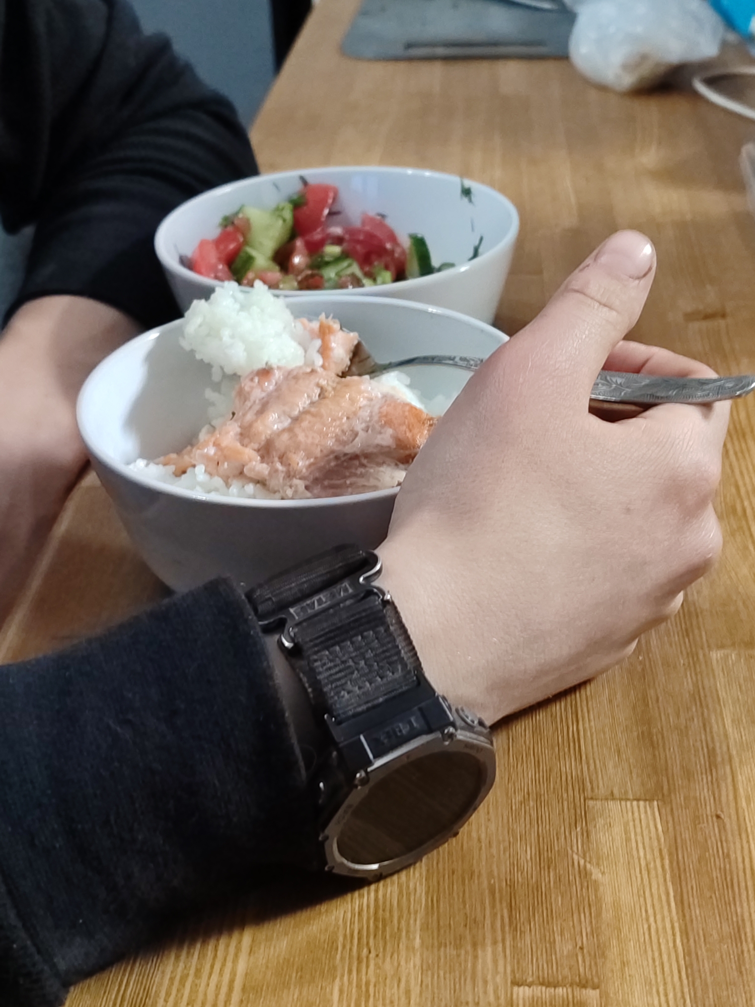 A close-up captures a person eating a meal at a wooden table. Two bowls are visible, one containing rice and salmon, and the other a salad with tomatoes and cucumbers. The person holds a spoon in their right hand. A black watch with a nylon strap is on the person's left wrist. The background includes an out-of-focus kitchen knife and a plastic bag on the table. There is no information that would confirm that this image was taken in the city of Chaikovsky, Russia.