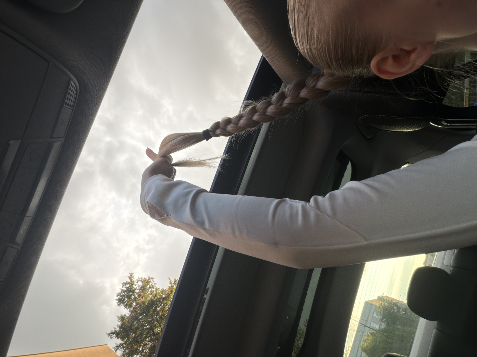 A low-angle shot taken from inside a vehicle, possibly a car, shows a person with blonde hair styled in a braid, holding the end of the braid up in their right hand. The braid extends from the top right corner of the frame towards the left, terminating near the left edge. The person is wearing a long-sleeved white shirt.
The background is a cloudy sky visible through an open roof or sunroof of the vehicle. Partially visible through the side windows are trees and buildings, suggesting an urban or suburban environment.
The interior of the vehicle is visible, including parts of the ceiling, a car seat, and window frames, all in dark tones. A control panel is seen on the top left corner.
