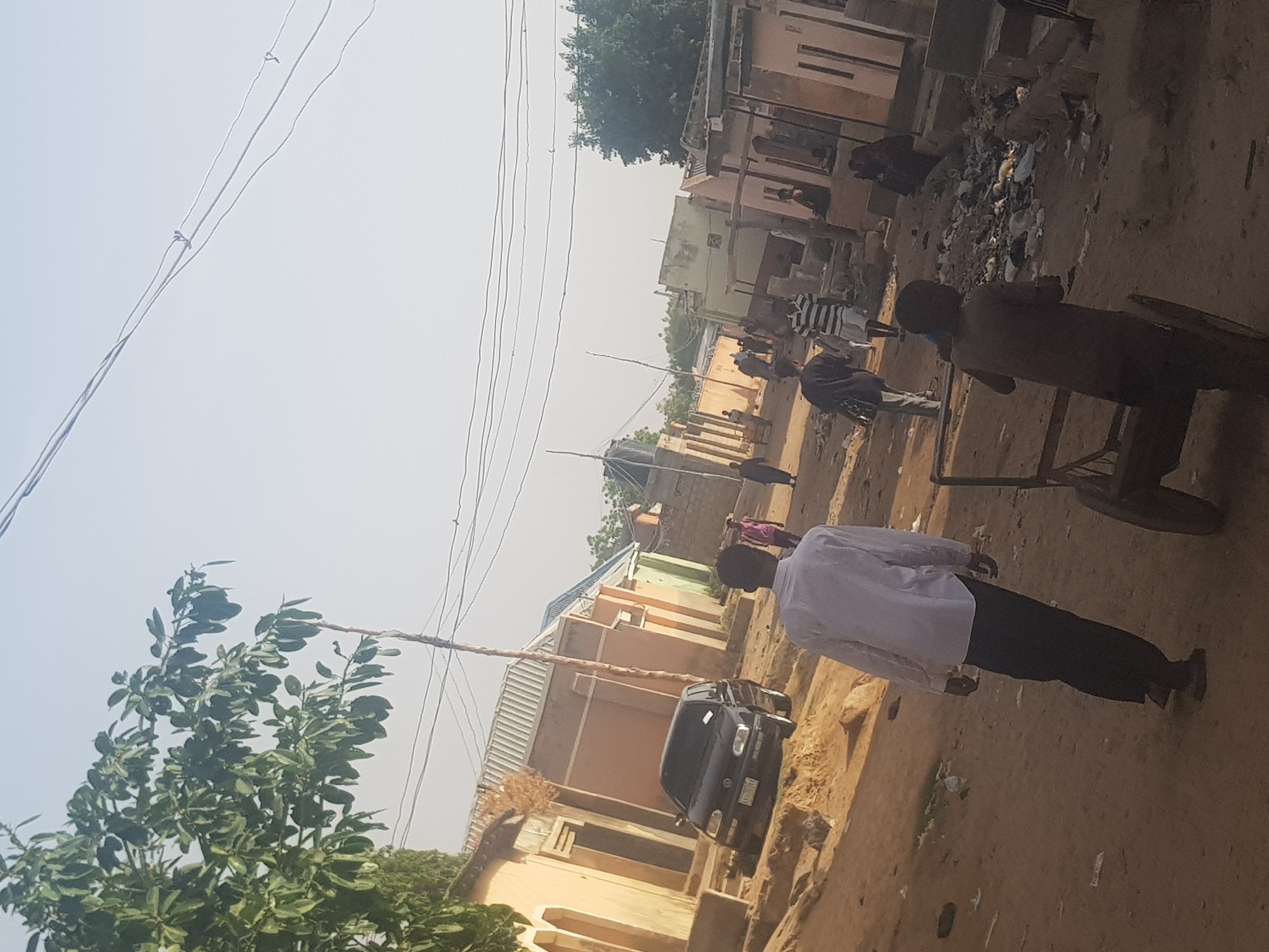 The image depicts a street scene in Dutsinma B, Nigeria. The environment is dusty and sunlit. Several people are present, some walking and others standing near what appears to be a pile of refuse. A man wearing a white shirt and dark pants stands in the foreground. Behind him, a person is pushing a wheelbarrow. A black car is parked along the side of the street in front of buildings constructed of light-colored materials. Power lines run overhead. Trees and other vegetation are visible on the left side of the image.