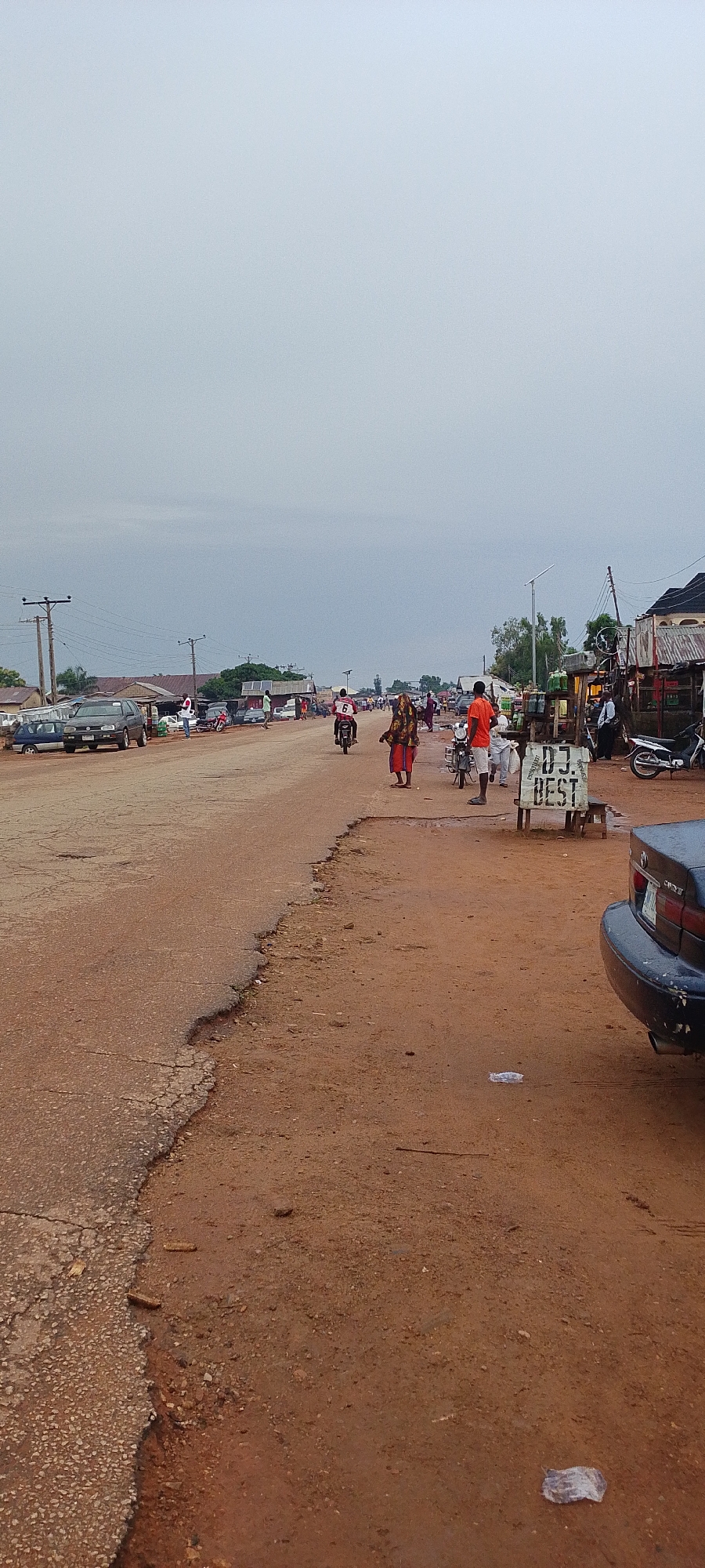 The image depicts a dusty road in Gumel, Nigeria, under an overcast sky, suggesting it is either early morning or late afternoon. The road is in disrepair, with a significant eroded section on the right side of the frame. A few vehicles, including cars and motorcycles, are present on the road and parked along its edges, indicating it's a functional route or marketplace.
Several people are visible, engaged in various activities. On the left, cars are parked, and some individuals appear to be near stalls or shops lining the road. In the center, a person on a motorcycle, wearing a red shirt with the number 6 on the back, rides along the road. To the right of the motorcycle rider, a woman in a colorful patterned dress and headscarf is walking. Further to the right, a man in an orange t-shirt stands near a roadside stall. Behind him, another man stands near a motorcycle.
A notable object in the foreground on the right is a signpost with the text "D.J. BEST" clearly visible. This suggests the presence of entertainment services in the area. The overall atmosphere appears to be that of a typical day in a Nigerian town, with commerce and daily life unfolding along the road, despite the signs of wear and tear on the infrastructure. The weather is cloudy, contributing to a subdued lighting condition.