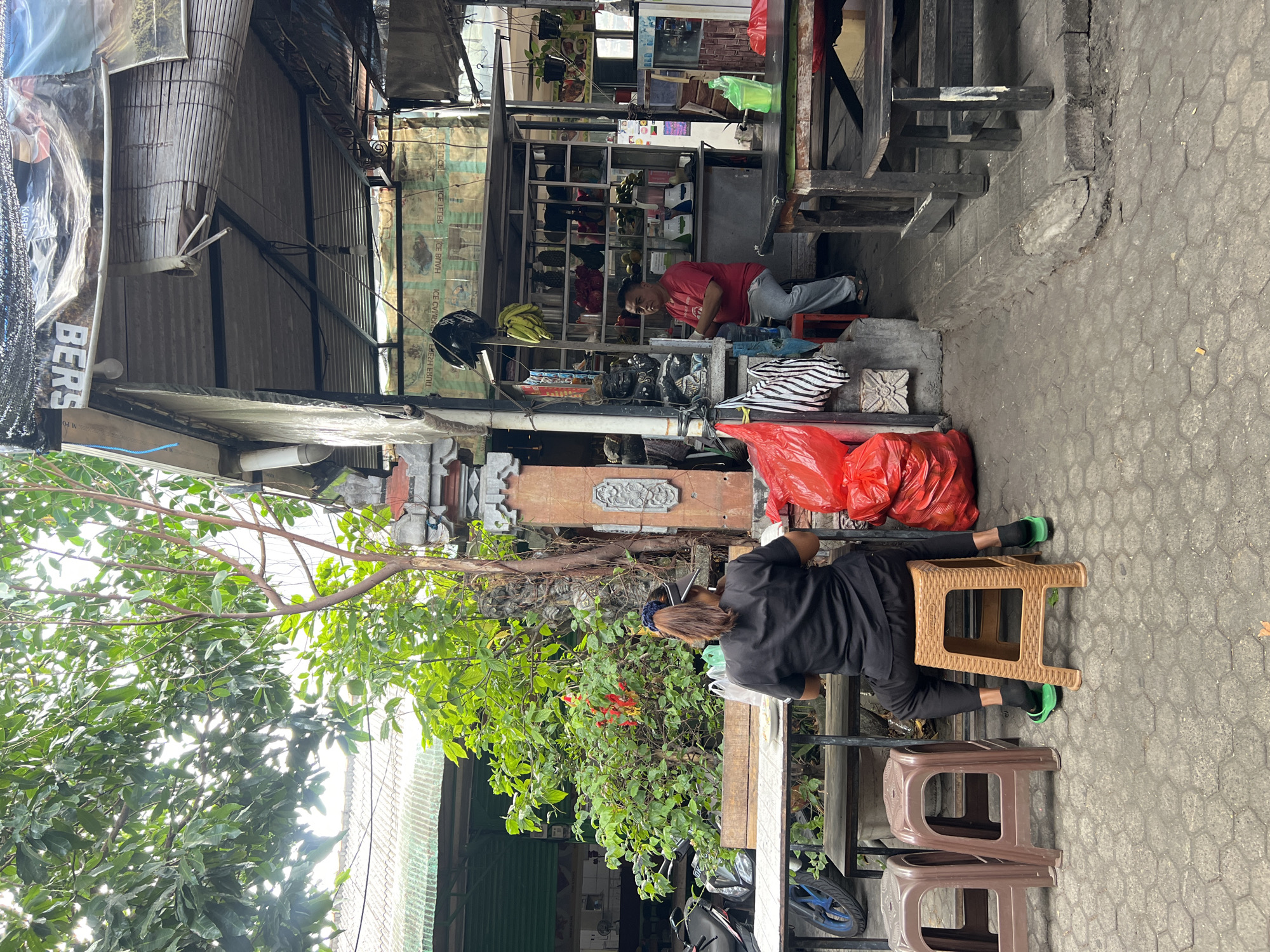 A street scene in Kuta, Indonesia depicts an outdoor food stall or juice bar. In the foreground, a person wearing a black shirt and pants is seated on a brown plastic stool, facing away from the camera. To their left are two identical brown plastic stools. A table sits in front of the seated person, with a small white bowl on it. To the right of the table is a large pile of red plastic bags.
In the middle ground, a man in a red t-shirt and gray pants sits on a red stool, facing the camera. He has his arms crossed. Behind him, the stall is stocked with various fruits and beverages, including pineapples, dragon fruits, avocados, and bottled drinks. There are blenders and other kitchen equipment visible. A motorcycle is parked to the left of the scene.
The environment is an outdoor setting with a tiled ground. Overhead, a corrugated metal roof is supported by metal posts, covered in some areas by tarps. Green foliage from trees and plants surrounds the stall. The overall impression is of a casual, local establishment in a busy area.
