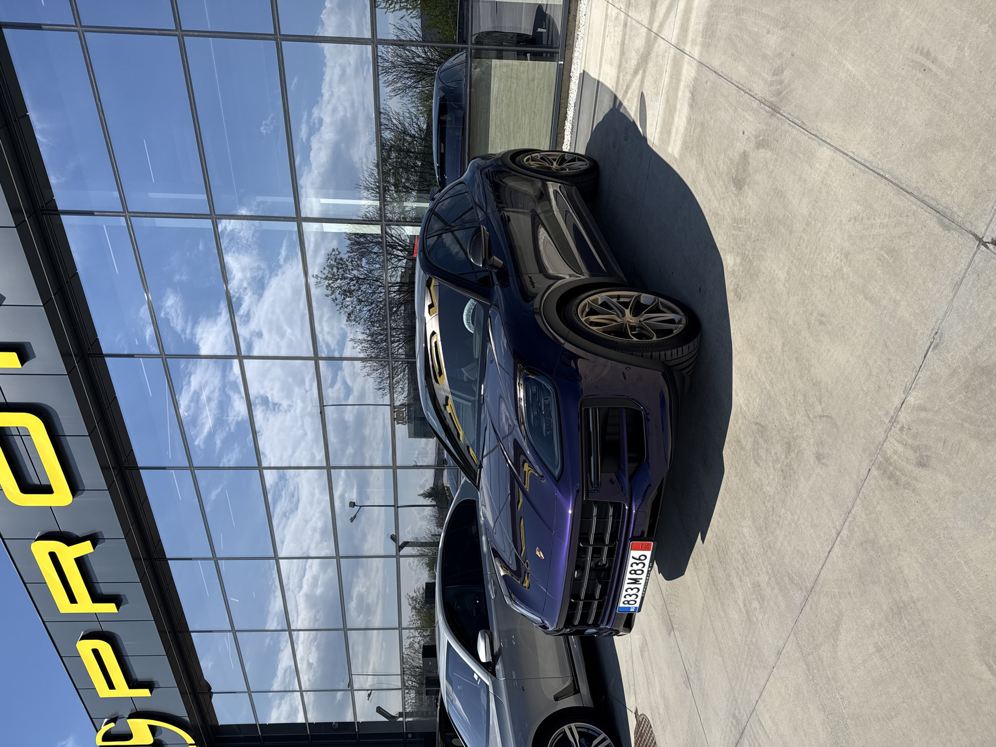 The photograph captures a deep purple Porsche Macan SUV parked on a concrete surface in front of a modern building. The car features distinctive golden-bronze multi-spoke wheels and has a Bulgarian license plate on its front bumper, displaying "BG 833 M 836" with "SOFIA" visible in smaller text below the registration numbers. Another dark-colored vehicle is partially visible behind and to the side of the purple Macan.
The building's facade is composed of large, reflective glass panels, which prominently mirror the clear blue sky with white, fluffy clouds, as well as bare tree branches. The reflections create a dynamic backdrop, intertwining the exterior environment with the building's surface. Part of a large yellow sign, displaying the letters "P O R", is visible on the building's exterior, strongly suggesting a Porsche dealership.
The scene is illuminated by bright daylight, casting long, sharp shadows from the vehicles and unseen objects onto the concrete pavement. The time of day appears to be midday or early afternoon, indicative of strong sunlight. The overall impression is that of a car showroom or dealership on a sunny day in Bulgaria. There are no people visible in the image, and no specific activity is taking place beyond the static display of the vehicles.