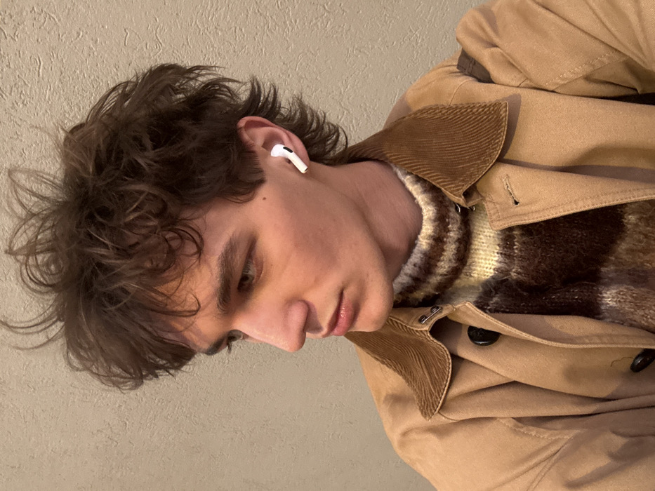 A young man with curly brown hair and an AirPod in his left ear looks downward. He wears a brown and beige striped sweater under a tan coat with a corduroy collar. The coat has black buttons and appears to be made of canvas or a similar durable material. The background is a plain, light beige wall.