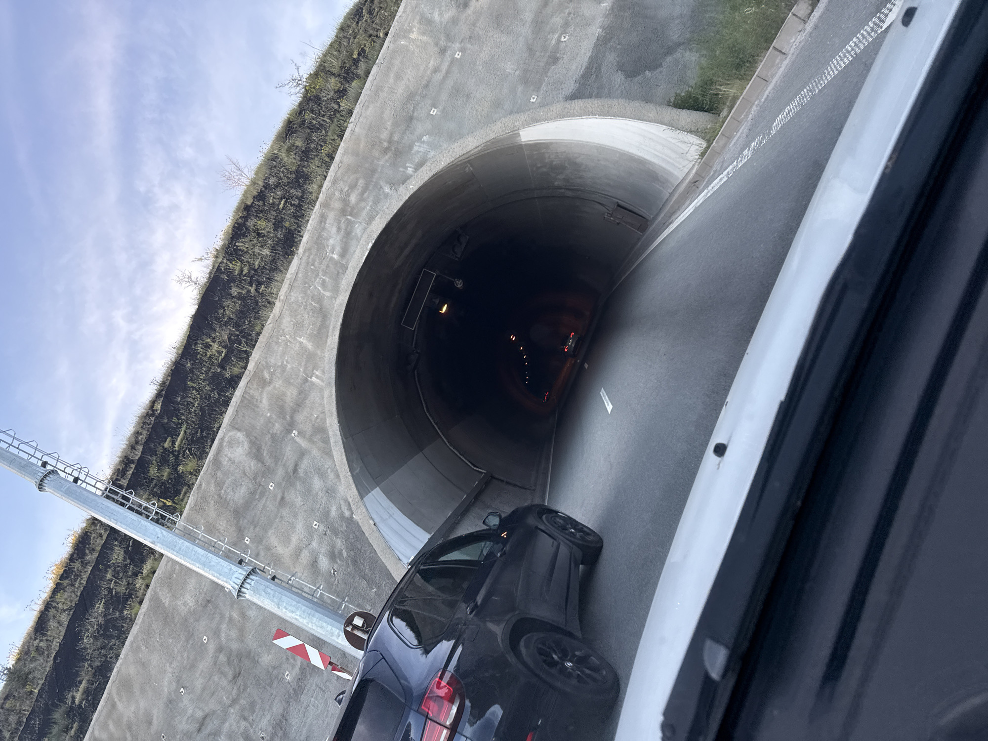 The image shows the entrance to a tunnel on a sunny day. A black SUV is parked to the left of the tunnel entrance, facing towards it. The tunnel is arched and made of concrete. There is a metal pole with a ladder on the left side of the tunnel entrance, and a red and white striped sign attached to it. The road leading into the tunnel is asphalt and has white lane markings. The surrounding area is hilly and covered with vegetation. The sky is blue with some clouds. The image appears to be taken from inside another vehicle, as a portion of a white car's hood is visible in the foreground. No people are clearly visible in the image.