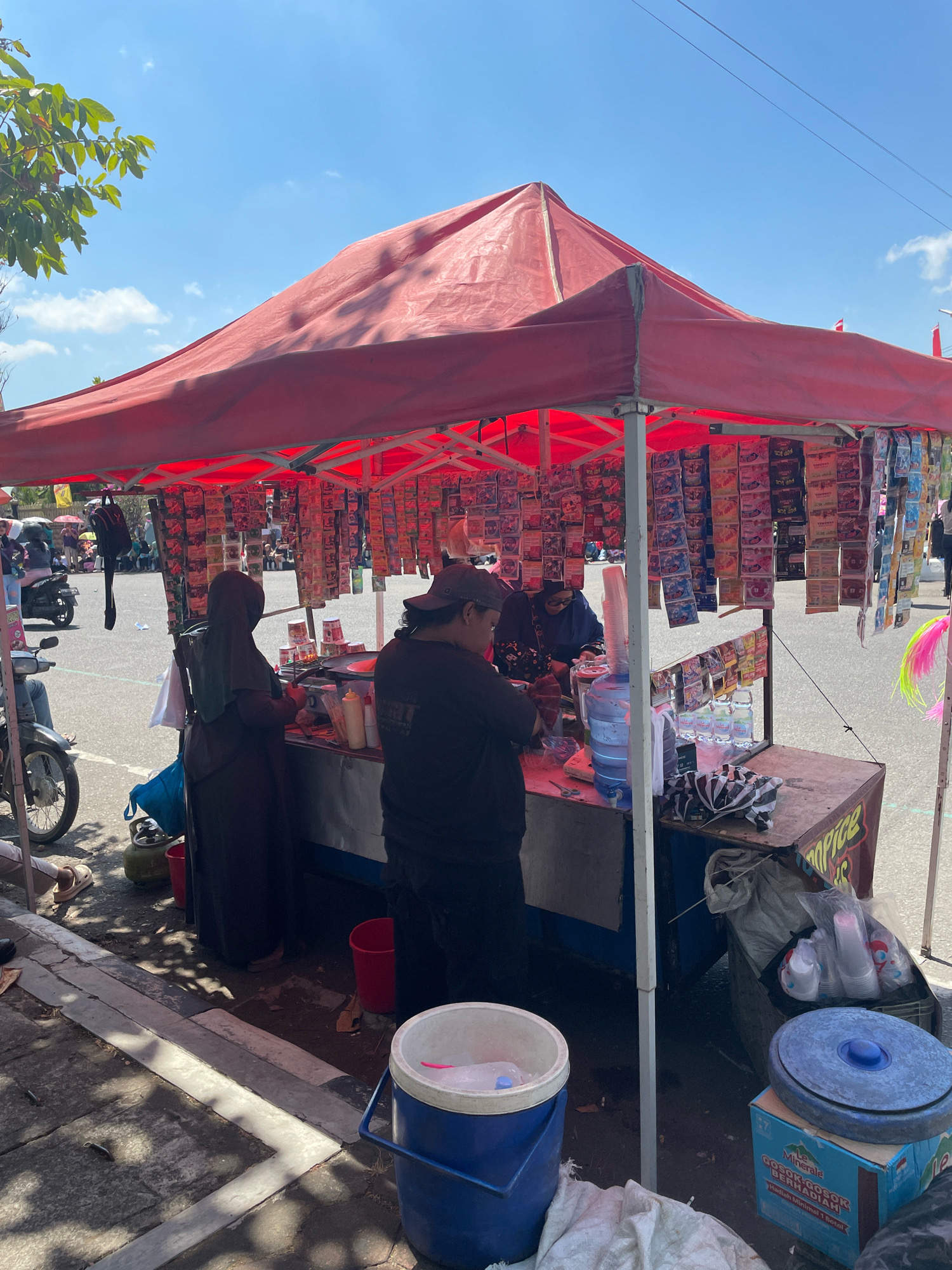 A street vendor operates a food stall under a red tent on a sunny day in Purworejo, Indonesia. Two individuals, one in a dark hijab and the other wearing a cap, are attending to customers or preparing food behind the counter. The stall is laden with numerous small packets of snacks and drinks hanging from the tent's support structure. A large blue water cooler sits on the counter, along with bottles of water, cups, and condiments. In the foreground, a blue cooler bucket is visible on the ground, next to a green gas cylinder. Motorcycles and a few other people can be seen in the background, suggesting a busy outdoor marketplace or street. The ground is paved with concrete and asphalt, with some debris scattered around the stall. The overall scene depicts a typical street-side commercial activity in Indonesia.