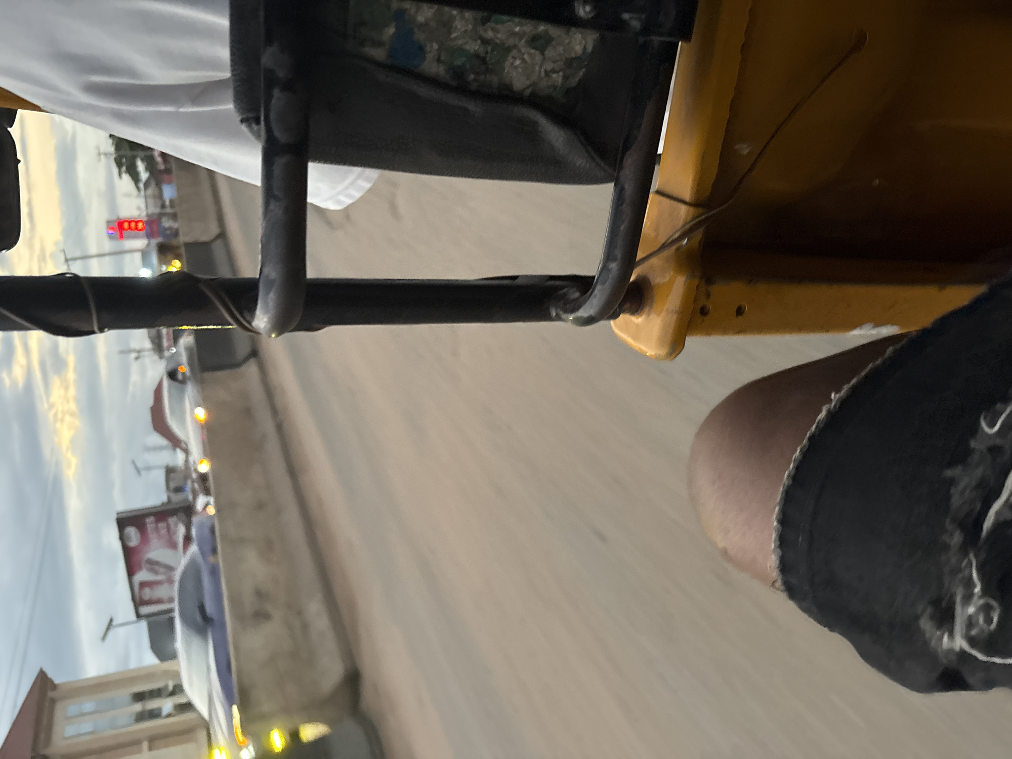 The image captures a first-person perspective from a yellow auto-rickshaw on a street in Onitsha, Nigeria. The foreground shows a portion of the passenger's right leg, clad in black, distressed denim shorts. The passenger is seated in the auto-rickshaw, indicated by the yellow frame and a black seat visible to the right. In the middle ground, a concrete barrier separates the auto-rickshaw's lane from oncoming traffic. Several cars are visible, including a blue sports car and a white car, with their headlights illuminated. A billboard with red and white lettering is seen behind the blue car. The background reveals a cloudy sky with hints of sunset and distant buildings, including a red neon sign with the number "100". The overall scene suggests movement along a busy urban road, possibly during dusk.