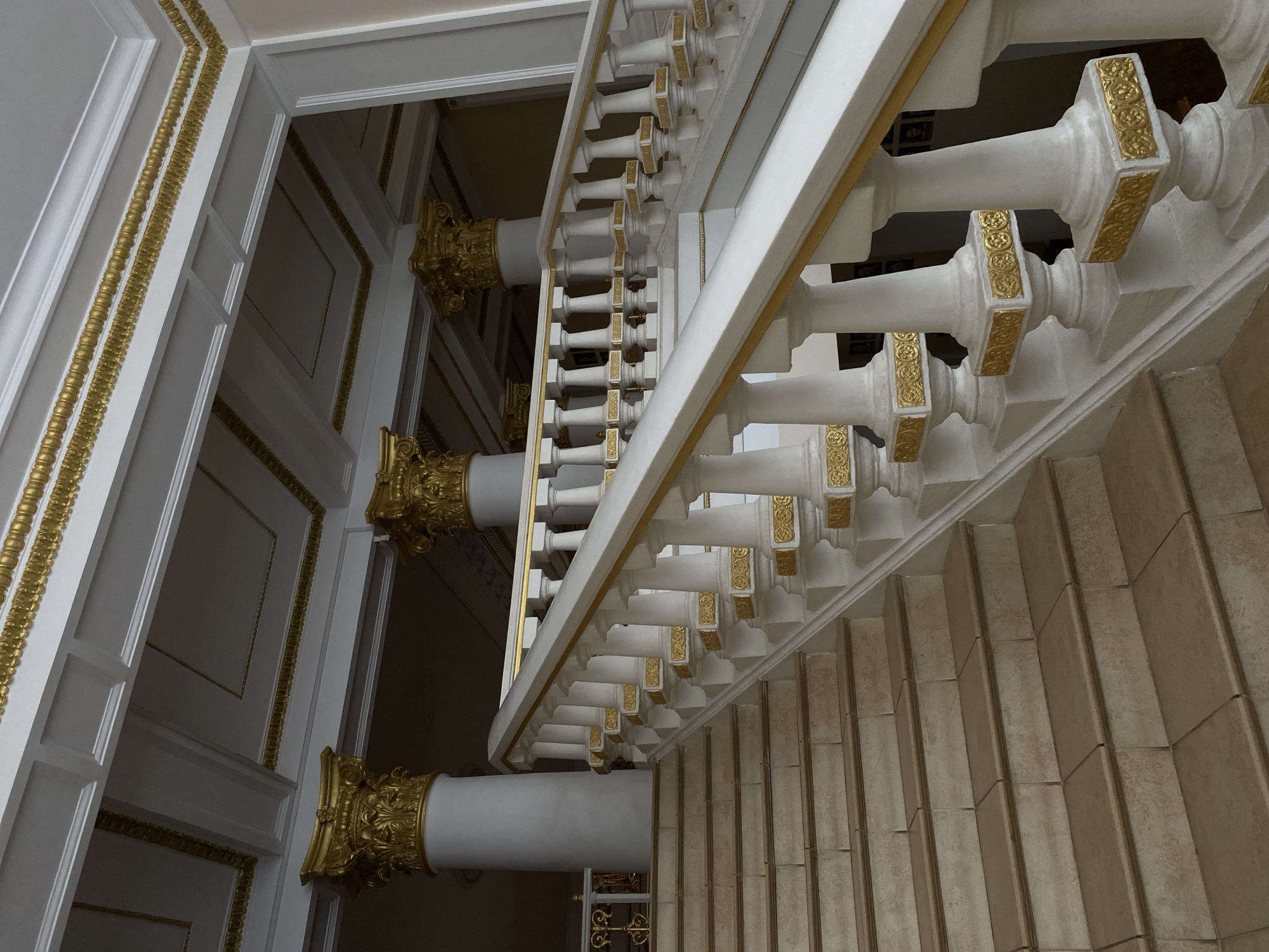 The image captures the view looking down a set of stairs, likely within a building in an unknown city. The stairs are constructed with light brown or beige material, possibly stone or tile, and extend out of frame downwards. On the right side of the stairs is a white balustrade with decorative gold accents on the posts. The balustrade runs along the length of the stairs. To the left of the staircase, are white columns with ornate gold capitals, creating a sense of grandeur. The wall is white, with recessed rectangular panels outlined with gold trim. The lighting is subdued, suggesting an interior space with controlled illumination. The composition of the image gives a sense of depth and height.