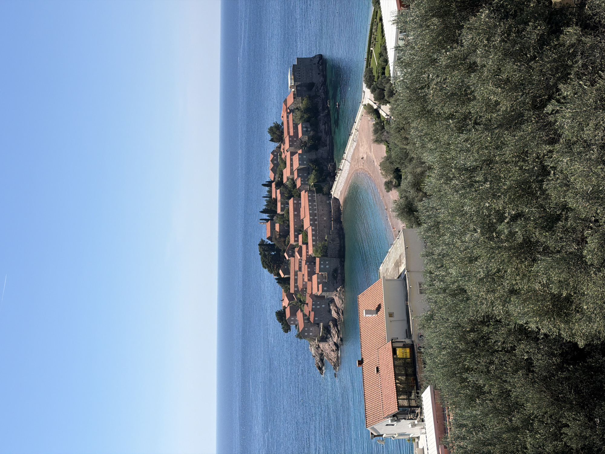 The image depicts Sveti Stefan, Montenegro, featuring an island with a cluster of buildings with red-tiled roofs connected to the mainland by a narrow isthmus. The Adriatic Sea surrounds the island, contrasting with the clear blue sky. Vegetation is visible along the mainland coastline. Buildings and trees partially obstruct the view on the right side of the image.