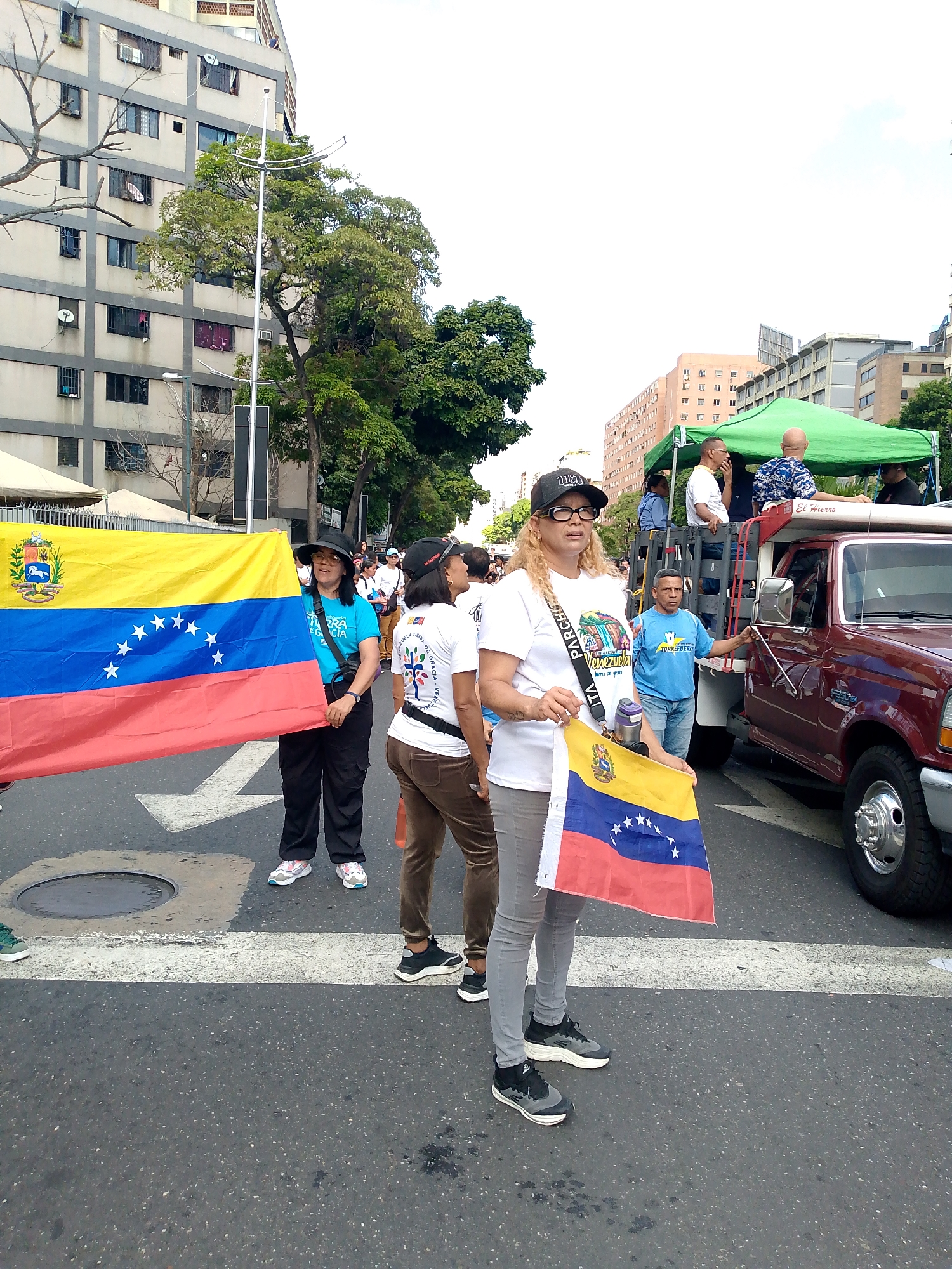This is a street-level, medium-shot photograph taken outdoors in what appears to be a city environment, likely Caracas, Venezuela, given the presence of the Venezuelan flag. The scene captures a street with a marked crosswalk in the foreground.
In the center of the image, a woman wearing a black baseball cap, glasses, and a white t-shirt with a graphic is holding a small Venezuelan flag. She is also wearing grey pants and black and white sneakers. To her left, a woman with dark hair and a black hat wears a bright blue t-shirt and black pants. She is holding a large Venezuelan flag horizontally. Behind them, other people are visible, some wearing t-shirts and caps.
To the right of the main subjects, a burgundy pickup truck is parked. The truck has a trailer bed with people in it. A green canopy is set up over the trailer bed. The background shows multi-story buildings and trees, typical of an urban setting. The lighting suggests it is daytime, with a slightly overcast sky. The overall impression is of a public gathering or event taking place on a street.