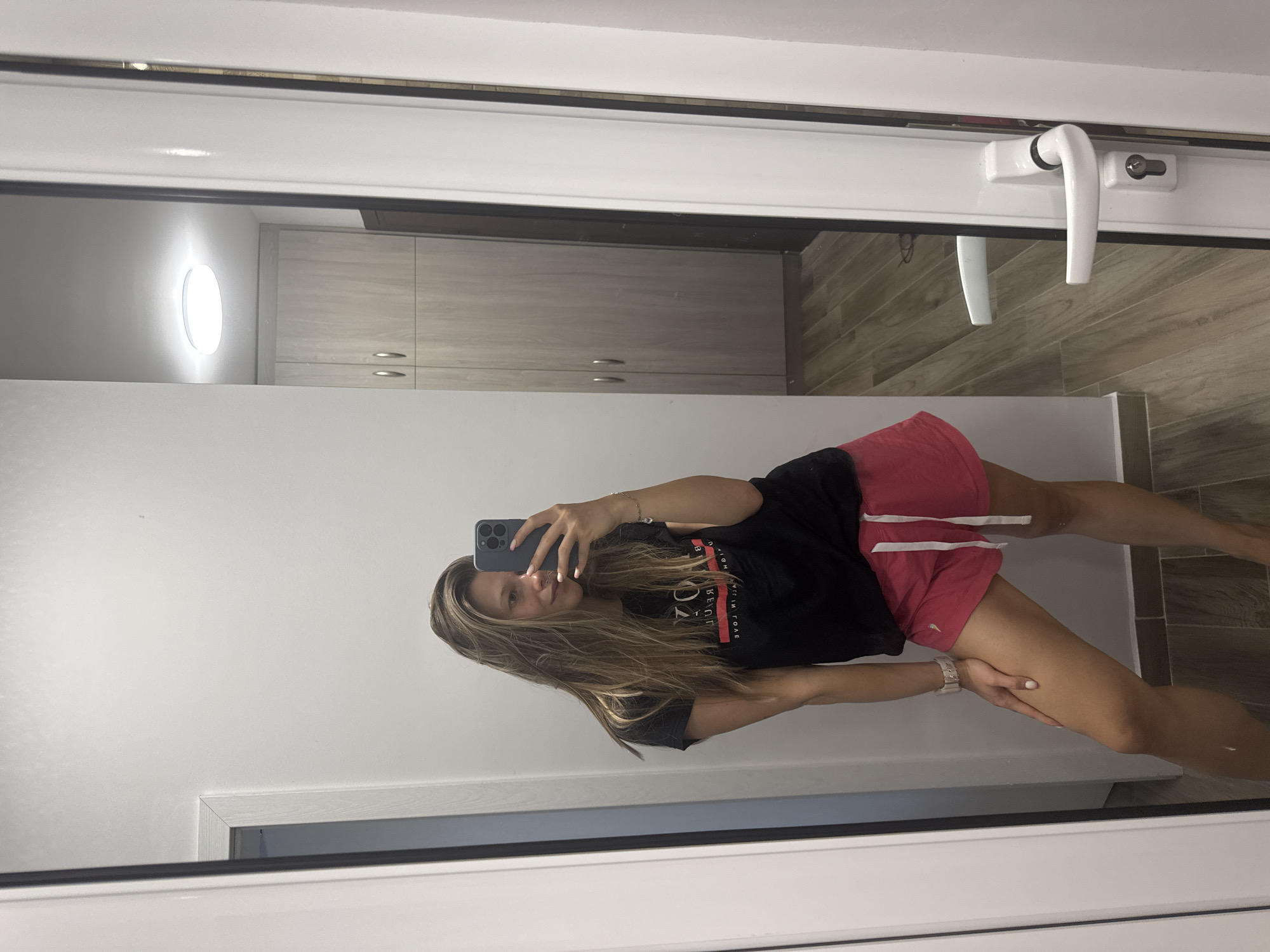A young woman with long, blonde hair is taking a selfie in a full-length mirror. She is wearing a black t-shirt with a red and white logo and pink shorts. Her left arm is resting on her left thigh, and her right hand is holding a smartphone. The mirror is in a room with white walls and a wooden floor. In the reflection, a door with a white handle and a keyhole can be seen, as well as a large wooden wardrobe and a circular ceiling light. There is no specific indication that this scene is in Sofia, Bulgaria.